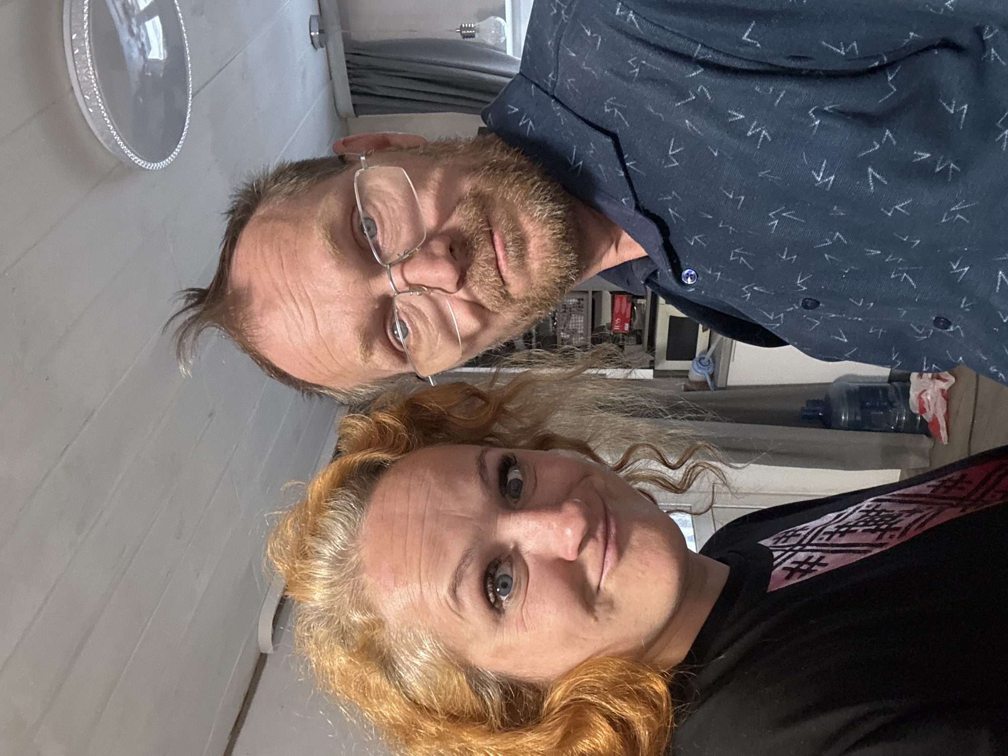 Two adults, a man and a woman, are captured in a close-up indoor shot, likely a selfie, in Chaikovsky, Russia. The image is oriented sideways, presenting the subjects horizontally.
The man, positioned on the left side of the frame (when correctly oriented), has light brown hair, a reddish-brown beard, and is wearing clear-framed glasses. He looks directly at the camera with a neutral expression. He is dressed in a dark blue button-up shirt adorned with a repeating pattern of small, light-colored geometric or arrow-like symbols. One of his cuff buttons is visible, appearing blue with a metallic center.
To his right (or below him in the sideways orientation) is a woman with long, curly, reddish-blonde hair. Her blue eyes are focused on the camera, and she has a subtle, faint smile. She wears a black top that features a distinctive red and black patterned design across the shoulder and upper chest area.
The background suggests a casual indoor setting. Behind the man's head, a decorative silver-rimmed tray or mirror is affixed to a white paneled wall. Further back, blurred elements include a large blue water dispenser bottle, dark curtains, and other indistinct household items. A bare lightbulb is also faintly visible in the upper background.
Both individuals appear relaxed as they pose for the photograph. There is no discernible text visible in the image.