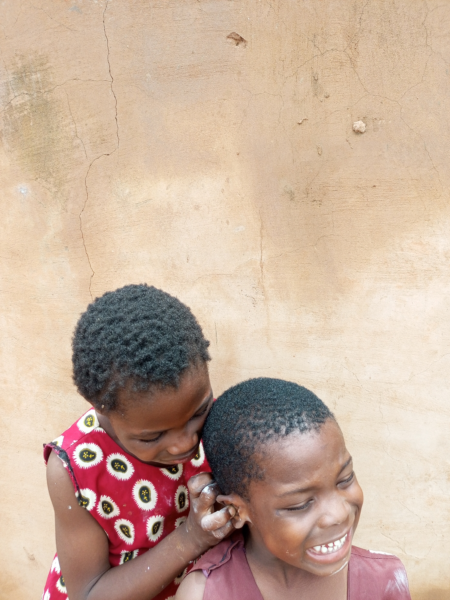 Two young children of African descent are positioned against a weathered, light brown stucco wall in Tempure, Nigeria. The child on the left, wearing a red dress with a white and black circular pattern, leans in and appears to be touching the ear of the child on the right. The child on the right is looking away with their eyes closed and mouth open, revealing their teeth in what seems to be a laugh or a grimace. Both children have dark, short, curly hair and dark skin. The wall behind them exhibits cracks and has a rough texture, indicative of age and exposure to the elements. The lighting suggests an outdoor setting during the daytime.