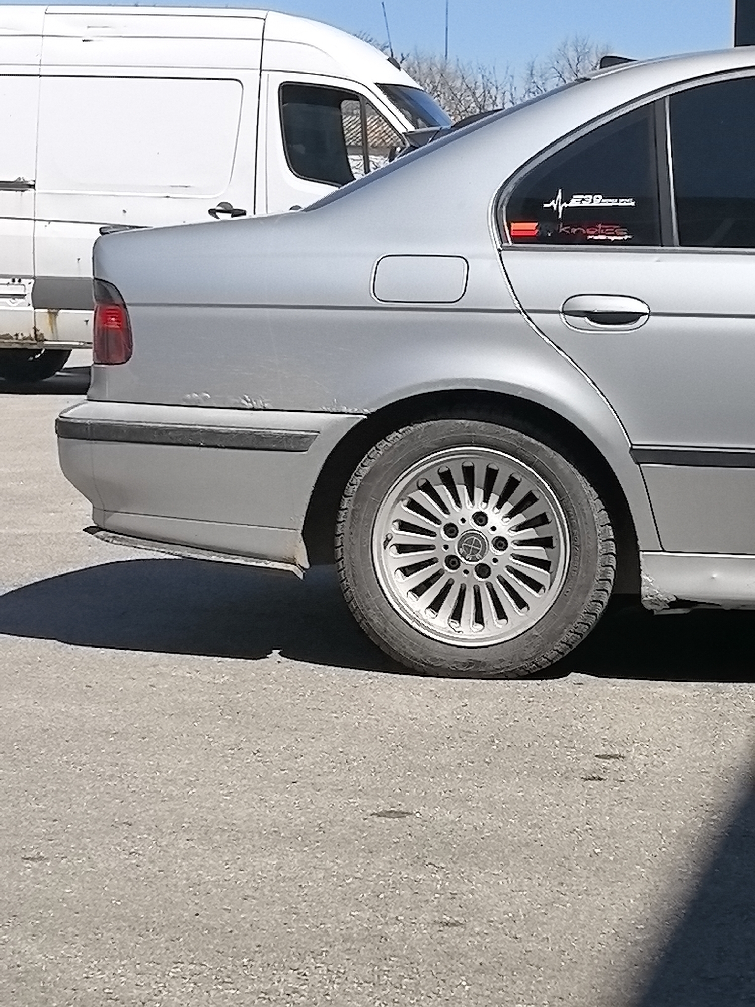 The image depicts the rear section of a silver BMW E39 sedan parked on asphalt. The car has alloy wheels and a sticker on the rear passenger window. A white van is partially visible behind the car. The location is Plovdiv, Bulgaria, but there is no evidence from the image.