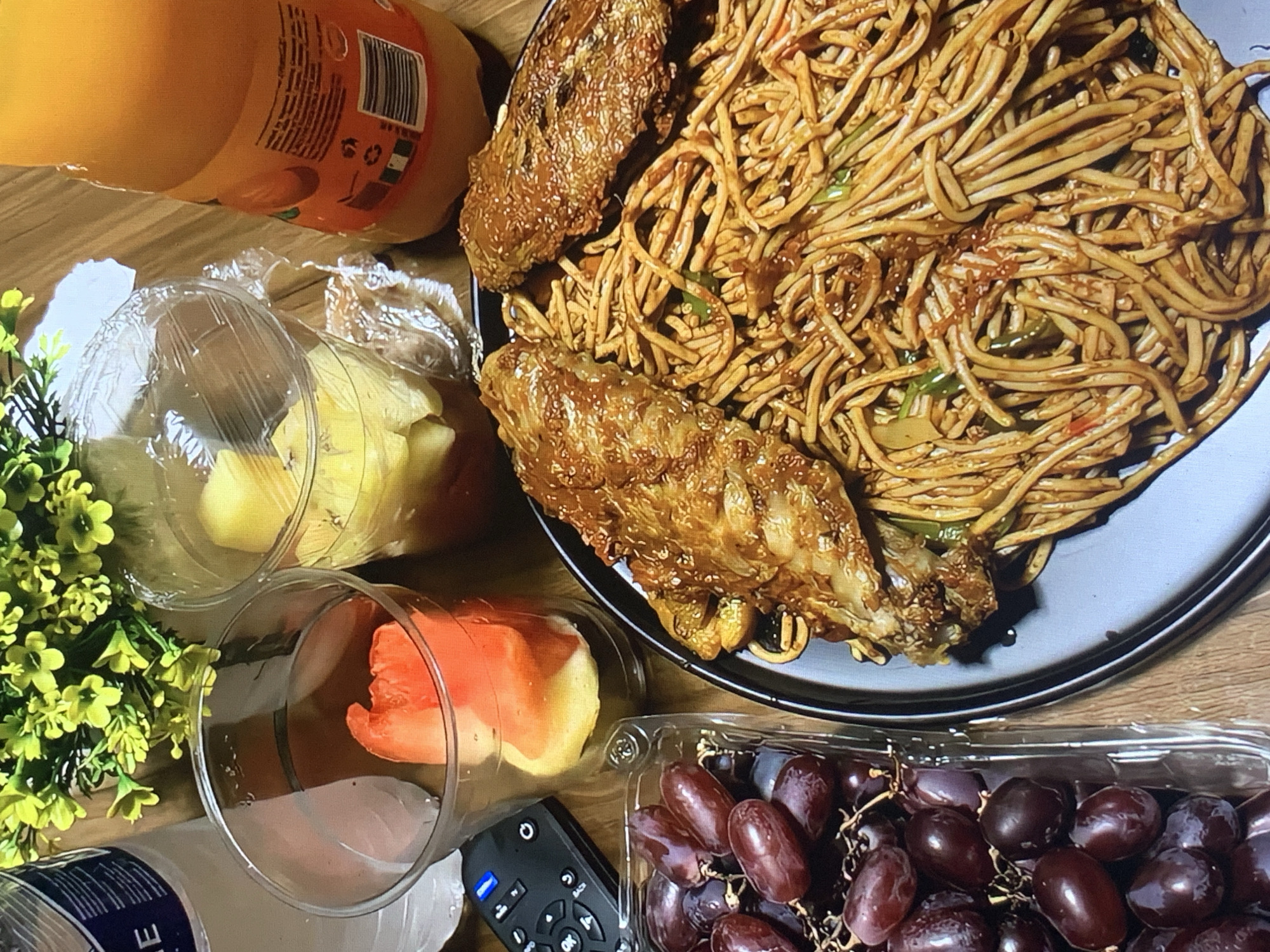 The image shows a wooden table cluttered with various food items. A black plate dominates the upper right of the frame, laden with spaghetti and two pieces of what appear to be fried chicken. To the left of the plate, there is a bottle of orange juice and two clear plastic cups, one containing yellow fruit and the other orange fruit. A small bouquet of artificial yellow flowers is positioned beside the cups. In the bottom right corner, a clear plastic container holds a bunch of dark red grapes. A black remote control is partially visible between the cup with orange fruit and the grapes. The location is not specified.