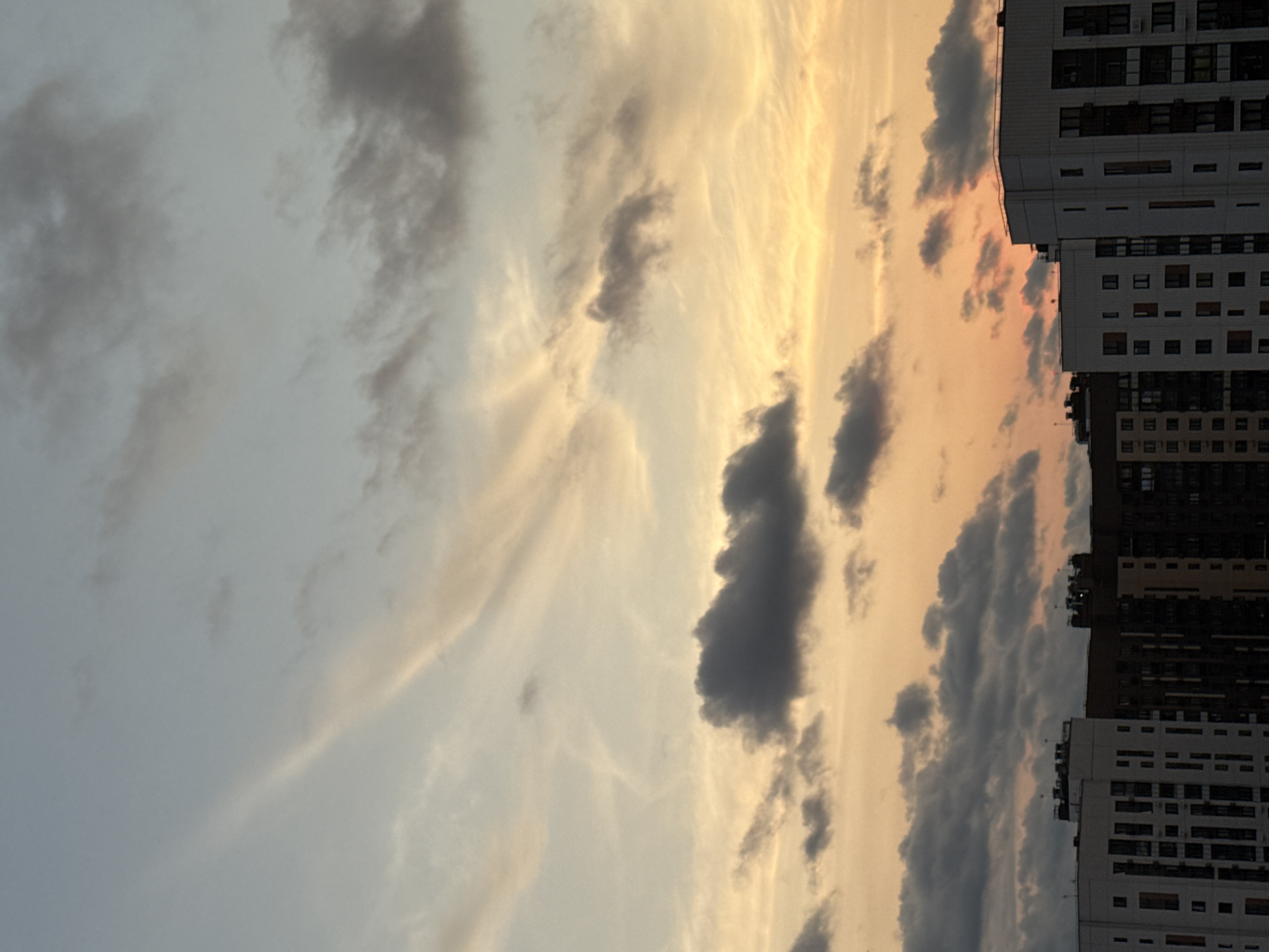 This is a vertical, medium shot photograph taken at dusk, looking upwards towards a sky filled with clouds and silhouetted apartment buildings. The sky displays a gradient of colors, transitioning from a pale blue at the top to warm oranges and yellows closer to the horizon. Wispy, light-colored clouds are scattered across the upper portion, while darker, more textured clouds are present in the middle and lower sections of the sky.
In the foreground, the tops of several multi-story apartment buildings are visible. These buildings appear to be modern, with a mix of white and darker facades, featuring numerous windows. The structures suggest a residential urban environment. The image was taken in the city of Lyubertsy, Russia. There are no people visible in the photograph. The overall scene depicts a cloudy sunset over an urban landscape.