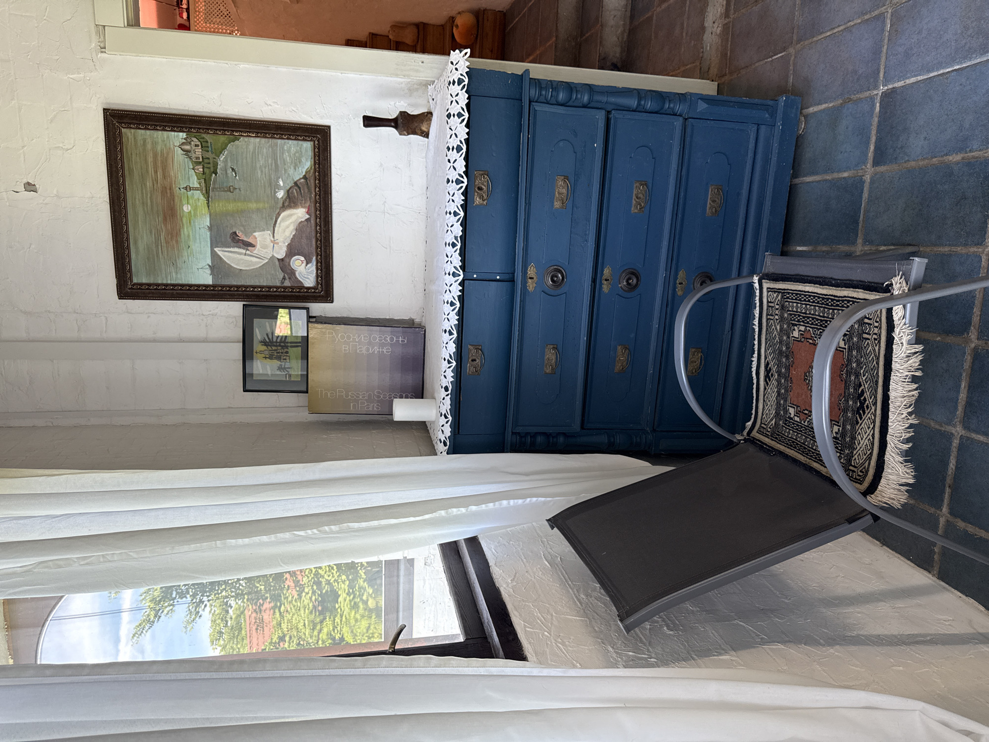 In Alagir, Russia, a corner of a room is depicted with a window on the left, covered by white curtains. Through the window, trees and a red-tiled roof are visible under a blue sky. A textured white wall is present. In the center of the image is a blue dresser with ornate brass drawer pulls. A white doily or tablecloth with an intricate pattern adorns the top of the dresser. On the dresser sits a white candle and two framed pictures, one larger with a painting of a woman on a rock by the sea and a smaller one with a depiction of a building. To the right of the dresser, a staircase with terracotta-colored risers and concrete steps leads upwards. Two pumpkins are visible on the staircase. In the foreground, a dark grey metal chair with a patterned rug on its seat is positioned in front of the dresser. The floor is covered with blue square tiles.