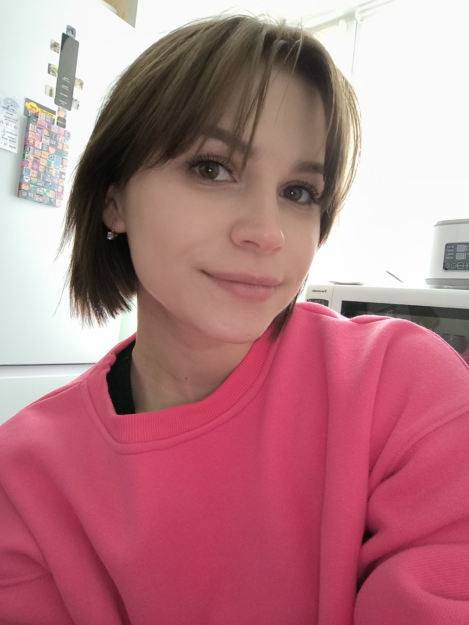 A medium close-up, eye-level indoor portrait features a young woman with short, straight brown hair and bangs, subtly smiling with light-colored eyes directed towards the viewer. She is wearing a vibrant, bright pink long-sleeved sweatshirt. A small, delicate silver hoop earring with a clear or light-colored stone is visible on her right ear.
The background suggests a home kitchen environment. To the left, a white refrigerator is partially visible, adorned with various magnets. One prominent, rectangular magnet displays numerous small, colorful, cartoonish faces arranged in a grid, with the word "WALLPAPER" faintly visible upside down on its top edge. Another metallic magnetic strip holds small photographs or notes, and a smaller magnet has illegible text.
On the right side of the frame, a white microwave oven is present, with the brand name "Panasonic" partially legible on its front panel. Above the microwave rests a smaller white kitchen appliance featuring a digital display showing "88:88" and several control buttons. The bright lighting suggests it is daytime. The woman's expression is calm and pleasant. The scene is set in Chaikovsky, Russia.