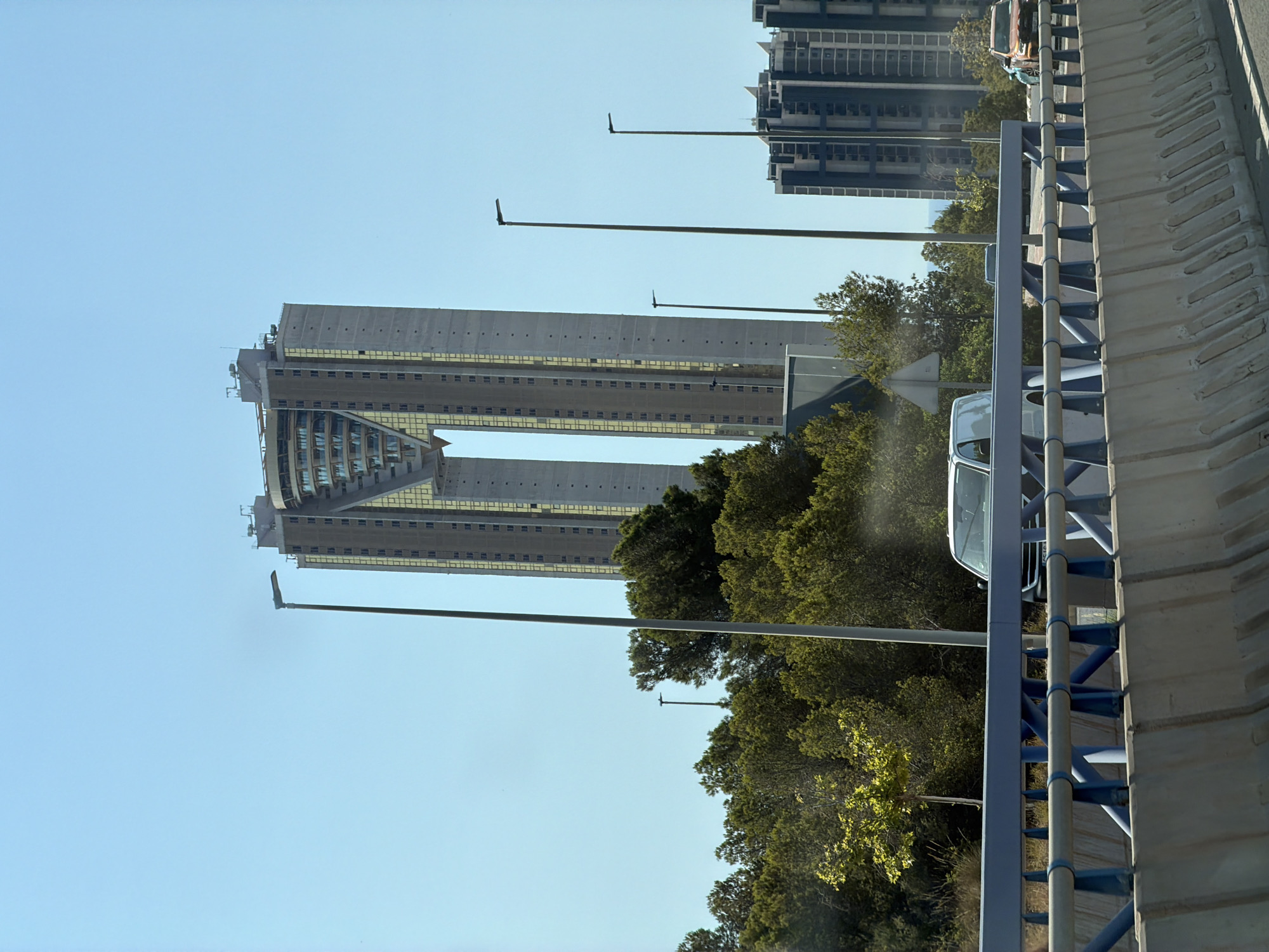 The image, taken from a highway overpass, shows a clear blue sky with the distinctive shape of the InTempo twin skyscrapers in Benidorm, Spain, dominating the background. The skyscrapers, notable for their inverted-cup design and large gap in the middle, are a light gray concrete color with yellow-tinted windows. Several streetlights with modern, minimalist designs are positioned in front of the buildings, along with a dense line of green trees.
In the mid-ground, the highway barrier, constructed of concrete with blue metal railings, runs horizontally across the frame. A white van and an orange car are visible on the highway. To the right, a cluster of more modern, dark-colored buildings with a grid-like facade are also present. The overall scene depicts a sunny day in a modern urban environment, likely a coastal city given Benidorm's location.