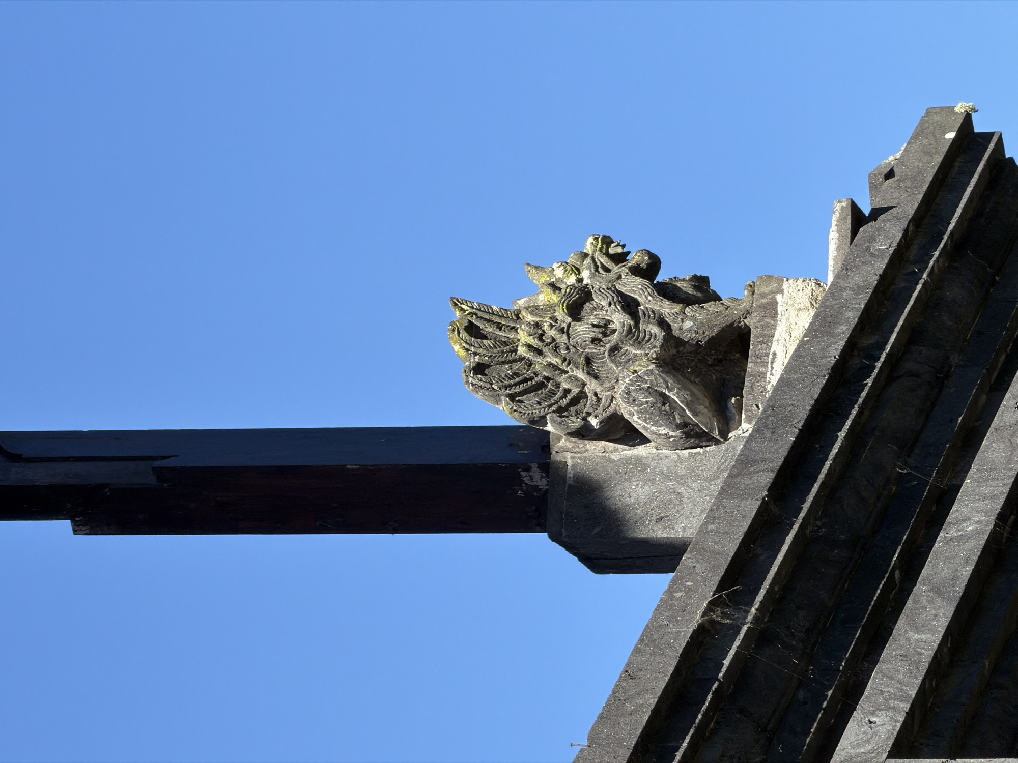 The image shows a close-up, low-angle view of intricate stone carvings adorning the exterior of a building against a clear blue sky. The carvings appear to depict a mythical creature or deity, with detailed features such as textured hair or mane, a distinct facial structure, and what seems to be decorative elements around its head, possibly resembling wings or elaborate headdress. The stone itself shows signs of age, with patches of moss or lichen adding to its weathered texture.
In the foreground and to the left, a dark, horizontal wooden beam juts out, casting a shadow on the stone structure below. To the right, a series of dark, angular, slatted stone elements rise diagonally, suggesting the roof or a decorative architectural feature of the building. The contrast between the rough, detailed stone carvings and the smooth, dark wood, along with the geometric lines of the architectural elements, creates a visually striking composition. Spiderwebs are visible on some of the stone surfaces, hinting at the environment and potentially the age of the structure.
The bright blue sky indicates it is daytime, likely with clear weather, as there are no clouds visible. The lighting suggests the sun is high, creating distinct shadows. The style of the carvings and the architectural elements suggest a location with a rich cultural or religious heritage, consistent with the provided location context of Baturiti, Indonesia, known for its Hindu temples and ornate stone work. No people or text are visible in the frame.
