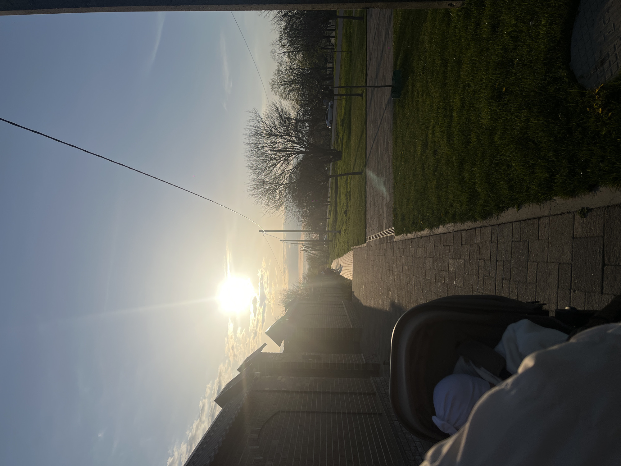 The image depicts an outdoor scene in Novopokrovskaya, Russia, captured from a low perspective, likely from someone pushing a stroller. The main subject in the immediate foreground is a dark-colored stroller, partially visible on the right, containing a baby or small child dressed in white clothing or a hat. The hand or arm of the person pushing the stroller is also visible, indicating an ongoing walk or outing.
The setting is a paved pathway made of brick-like blocks, stretching into the distance under a bright sky. To the left of the path, a series of brick buildings with dark, pitched roofs line the route. These structures cast long shadows across the path, suggesting a low angle of the sun. On the right side of the pathway, a vibrant green grassy area extends, populated with several bare, deciduous trees, which points to either late winter or early spring. A white car is partially visible parked further down near these trees.
The time of day is characterized by a low-lying sun, which is shining intensely and creating a strong lens flare and warm glow in the sky. This bright light and the long shadows strongly indicate either an early morning sunrise or a late afternoon/early evening sunset. The sky is predominantly clear with some scattered clouds, particularly around the sun. A thin, dark utility line or cable is visible stretching horizontally across the upper portion of the sky. No visible text is present in the image.