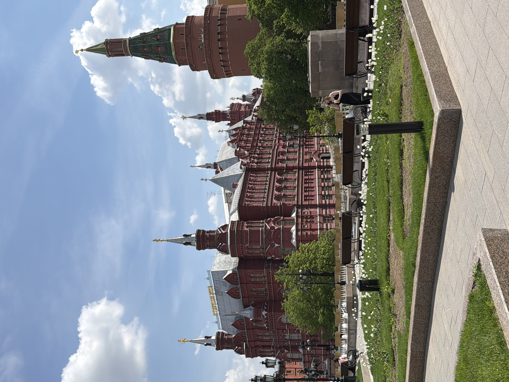 The image depicts a scene in Moscow, Russia, featuring the State Historical Museum and a section of the Kremlin Wall. The museum, a large red brick building with multiple spires topped with golden eagles, dominates the lower part of the frame. To its right, a segment of the Kremlin Wall, with its distinctive tower, rises into the sky. Fluffy white clouds dot the blue sky in the background.
A paved area extends from the bottom right, leading to a green strip of grass and flowerbeds interspersed with benches. One person is visible sitting on a bench. The lighting suggests it is daytime with sunny conditions.