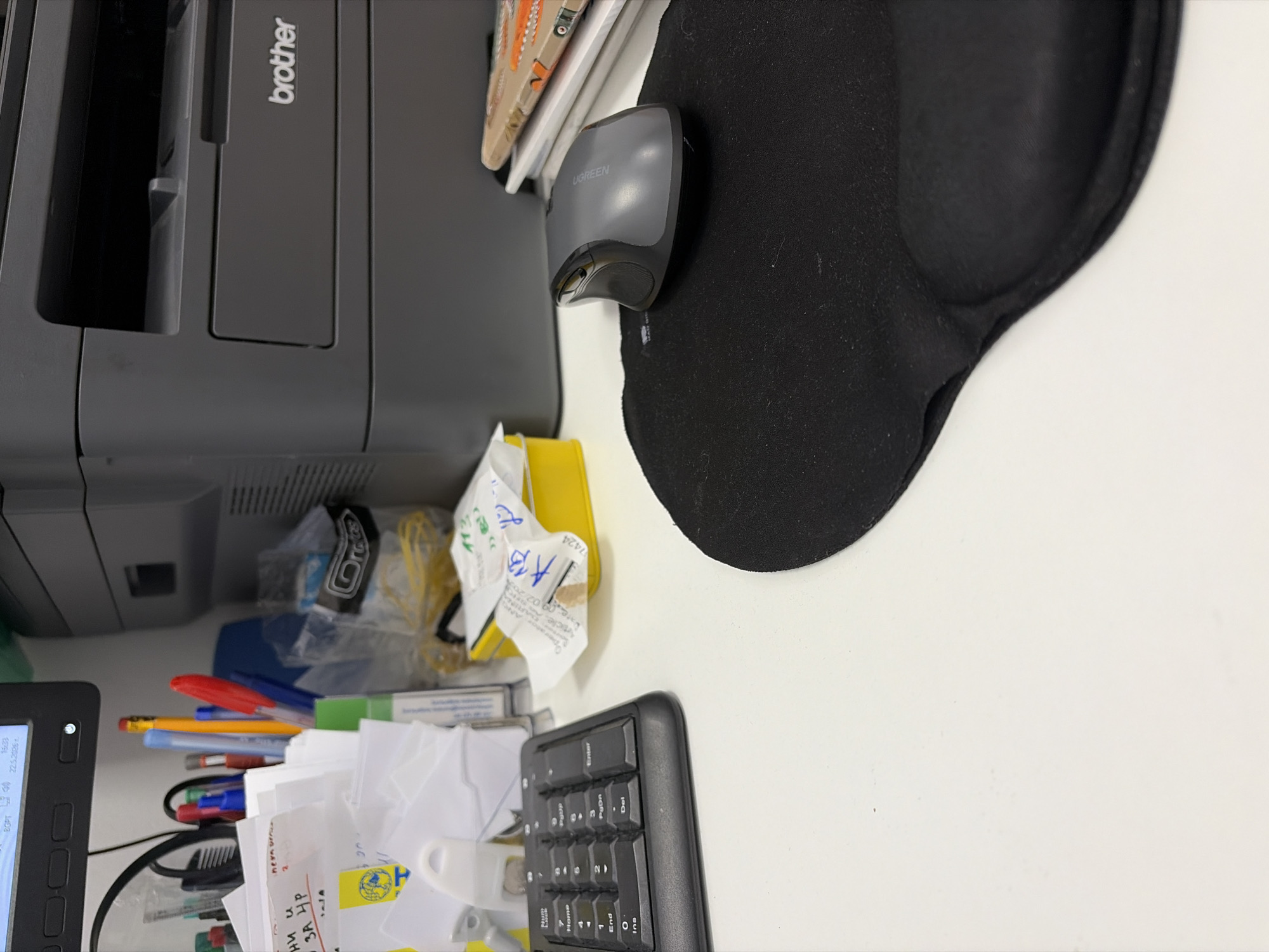 The image shows a cluttered desk. On the left is a gray Brother printer, next to a display screen, a bundle of pens and pencils, and crumpled paper. A yellow sticky note partially covers the paper. A black numeric keypad is on the bottom right. Above that is a black wrist rest and a gray Ugreen mouse. On the top left is a stack of books and papers. The desk is located in Stara Zagora, Bulgaria.