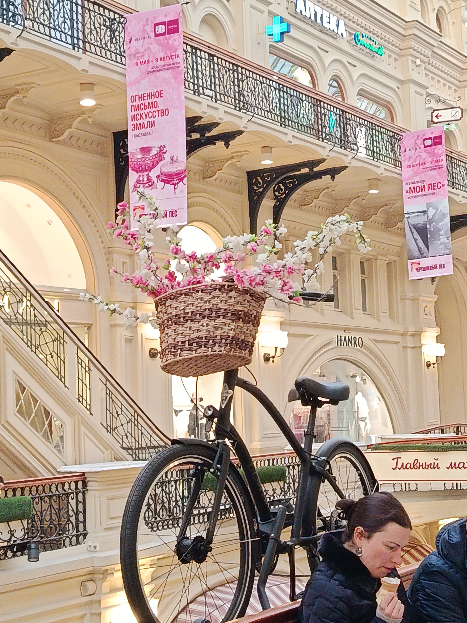 The image shows a scene inside a shopping mall. A black bicycle is prominently displayed with a wicker basket filled with white and pink flowers mounted on its front. In the background, there are multiple levels of the mall with white walls, arched doorways, and decorative railings. Banners are hanging from the upper levels, displaying text in Russian, including dates from May to August and the words "ОГНЕННОЕ ПИСЬМО. ИСКУССТВО ЭМАЛИ" (Fiery Letter. Enamel Art). A sign for "APTEKA" (Pharmacy) is also visible. On the ground level, people are seen in the lower right of the frame, and a sign for "Главный ма" (Main ma) is partially visible. The setting seems to be an upscale retail environment, possibly in a city with historical architecture.