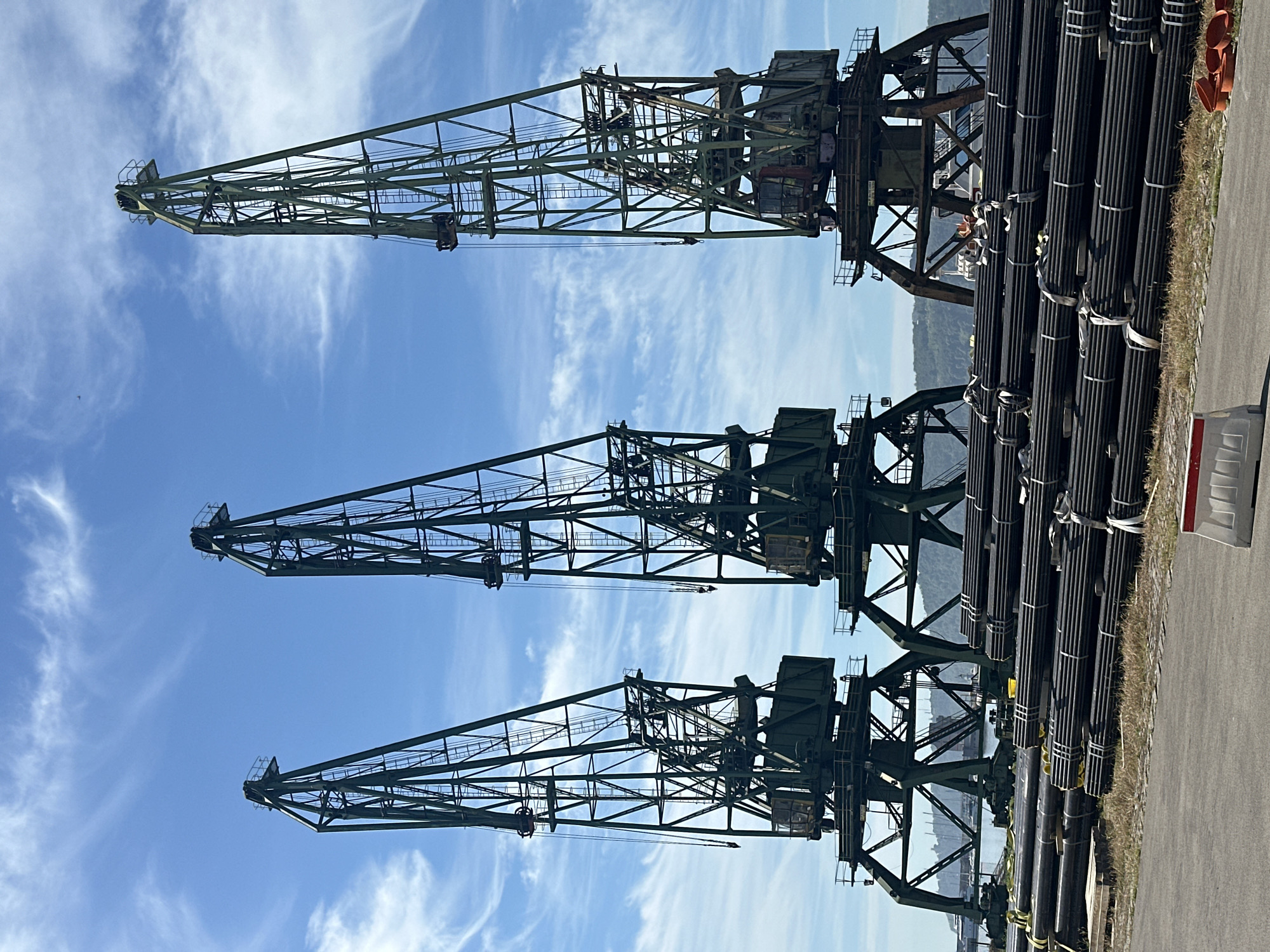 In an outdoor, industrial scene, three tall, dark green gantry cranes stand in a row against a bright blue sky with wispy white clouds. The cranes are constructed from a lattice of dark metal beams and have a robust, heavy-duty appearance. The leftmost crane is slightly offset from the other two, which are positioned closer together. In the foreground, a large stack of dark, cylindrical pipes, bound with white straps, lies horizontally. The pipes appear to be industrial in nature, possibly for construction or infrastructure. Behind the pipes and between the cranes, a body of water is visible, with a few boats and a larger vessel in the distance. Hills or mountains form a backdrop on the far side of the water. The ground in the immediate foreground is paved, with a strip of dry grass and a white and red barrier visible on the right. The overall impression is that of a working port or industrial storage facility.