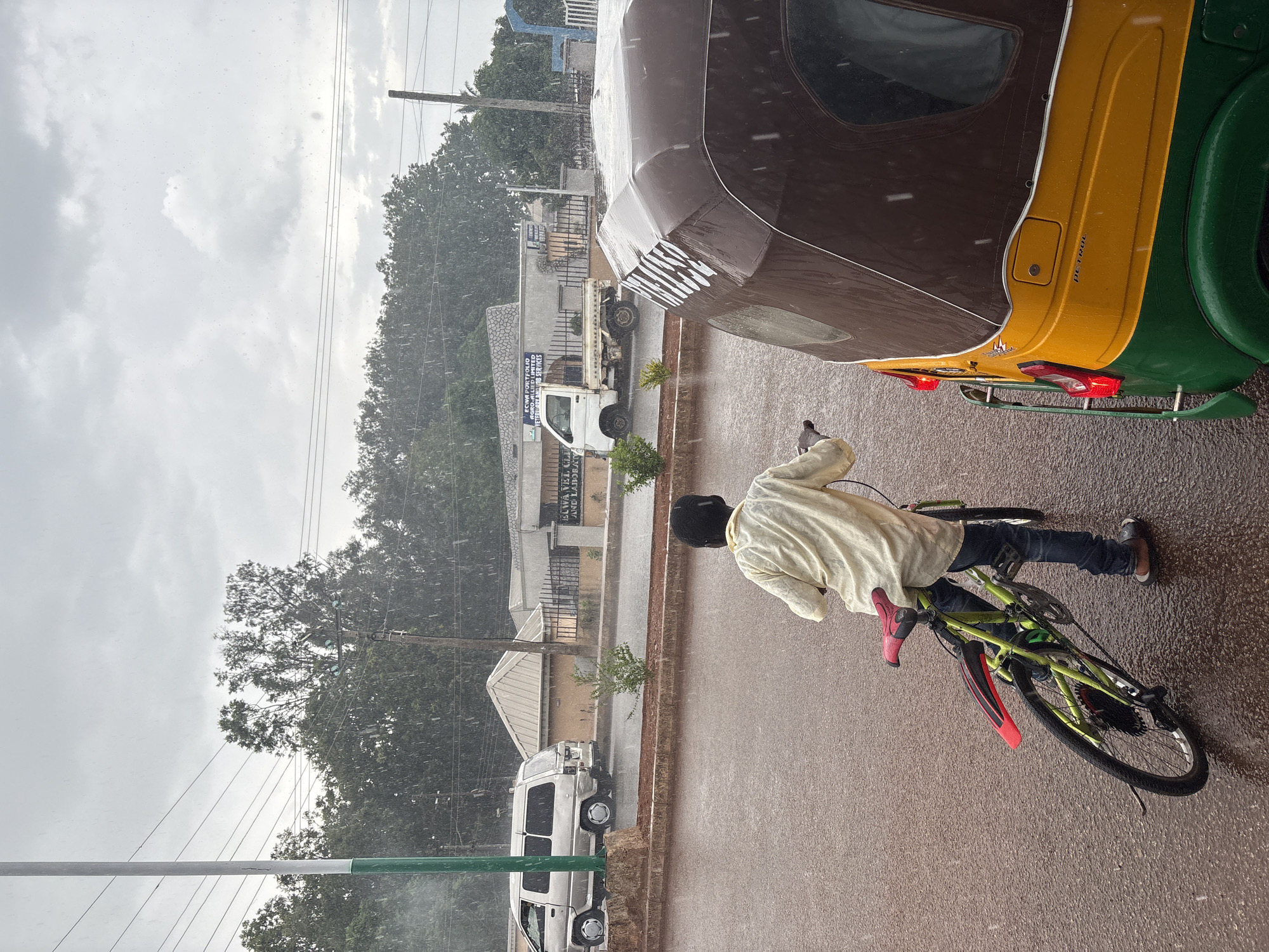The image shows a street scene in Bukuru, Nigeria during a heavy rain. A person in a light-colored top and dark pants is walking with a green and red bicycle on a wet road. A vehicle, possibly a three-wheeled "Keke Napep," is partially visible in the upper right. Other vehicles are parked alongside buildings on the left side of the street. Power lines and trees are visible in the background against a cloudy sky.