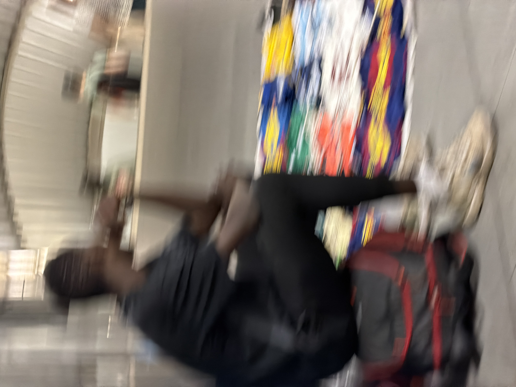 The image depicts a blurred scene that appears to be a market stall or a similar retail environment, possibly within Barcelona, Spain. A person, whose features are indistinct due to motion blur, is seated in the foreground. They are wearing a dark t-shirt and dark trousers. To their left, a dark bag with red accents is visible on the ground. Behind the person, a display of colorful fabric or clothing items is spread out, featuring prominent colors such as blue, yellow, and red, suggesting textiles or garments for sale. The background shows shelving units and a somewhat industrial-looking ceiling structure, hinting at an indoor marketplace or storage area. The overall impression is one of a busy, perhaps crowded, commercial setting.