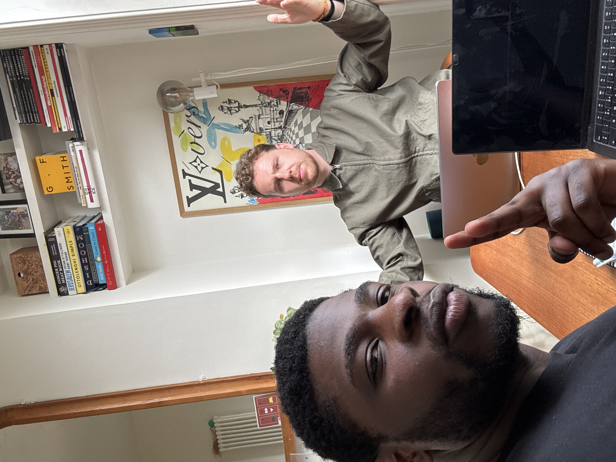Two men are depicted indoors, likely in a home or office setting, featuring a bookshelf and artwork. The image, taken from a slightly low angle, shows a Black man in the foreground, his right hand pointing towards a laptop on a wooden table. He has a neatly trimmed beard and is wearing a dark t-shirt.
Behind him, a White man with light brown, curly hair is visible, wearing an olive green jacket. He looks directly towards the camera with a neutral expression, his right arm raised. On the left side of the frame, a white bookshelf is filled with various books and magazines. Visible titles include "GINGER PIG MEAT BOOK," "JERUSALEM," "OTTOLENGHI SIMPLE," "SMITH," "DISHOOM," and "Wonderland." Other text mentions "connectivity," "Rick Stein," "Coast to Coast," and "PLATS PLAS."
A large framed artwork hangs on the wall behind the man in the background, featuring the word "L•vers" and the "LV" (Louis Vuitton) logo, intertwined with abstract cityscape elements and a checkered pattern. A vintage-style light fixture with an exposed bulb is mounted above the artwork. On the wooden table, an open black laptop is positioned, with a rose gold laptop or tablet partially visible beneath it. The pointing hand of the man in the foreground is near these devices.
The room appears well-lit, suggesting daytime. A radiator is visible in the bottom right corner of the original image, hinting at a European location, possibly London, United Kingdom. The overall scene suggests a casual interaction or moment captured within a personal living space.