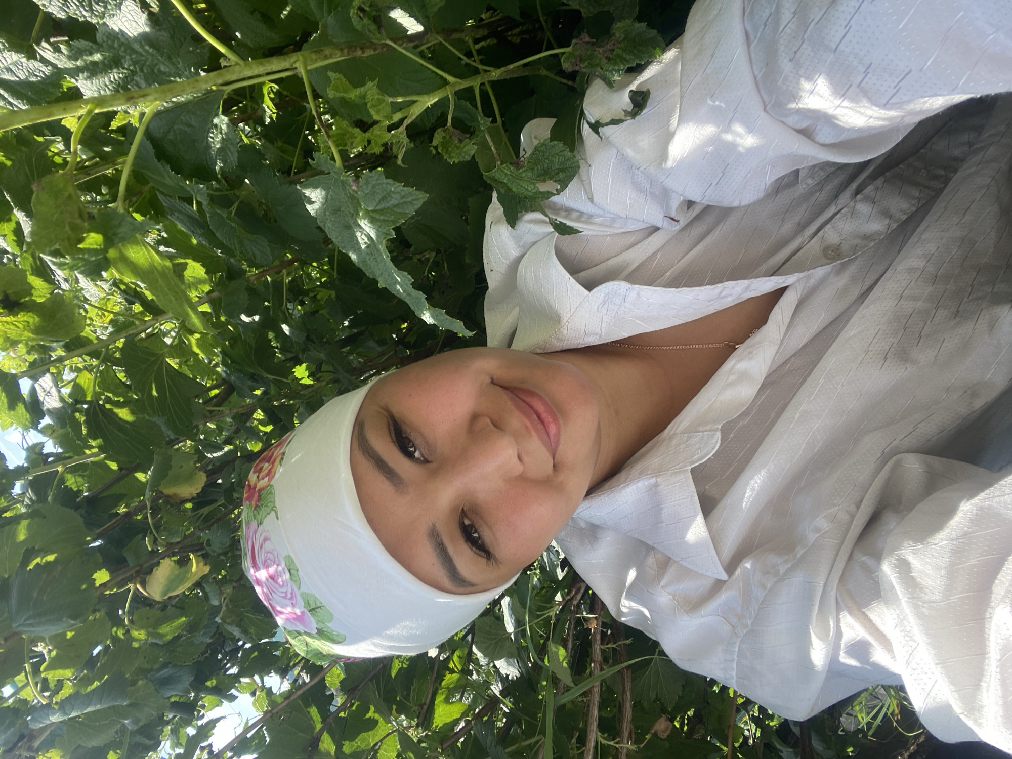 A medium close-up, self-portrait taken outdoors in Maloarslangulovo, Russia shows a young woman wearing a white headscarf with a floral print and a white button-down shirt. She is smiling and looking at the camera, with lush green foliage filling the background. The sunlight creates bright highlights on her clothing and skin.