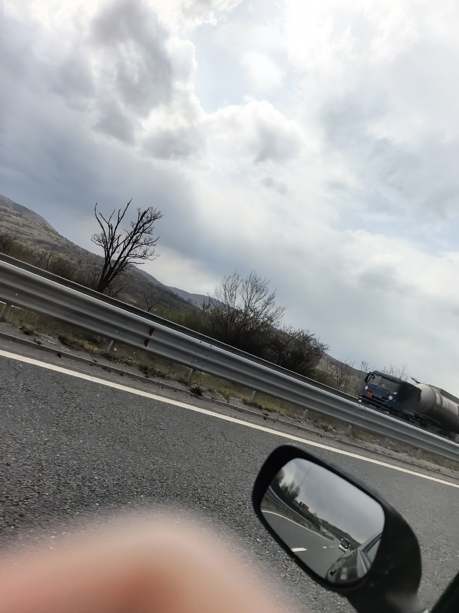 The image depicts a scene from a vehicle, likely a motorcycle, traveling on a multi-lane highway under an overcast sky. A prominent rearview mirror is visible in the bottom right corner, reflecting the road and other vehicles behind. A large finger, possibly belonging to the photographer, is partially visible in the bottom left.
In the midground, a silver metal guardrail runs parallel to the road. Beyond the guardrail, the landscape features bare trees, suggesting late autumn, winter, or early spring. In the distance, rolling hills or mountains are visible under the cloudy sky.
A dark blue truck with a tanker trailer is seen on the highway to the right, driving in the same direction as the viewer. The brand name "IVECO" is clearly visible on the front grille of the truck's cabin. The sky is predominantly gray with scattered lighter patches of clouds, indicating a cloudy or overcast day. There are no clear cues for the specific time of day beyond being daylight. The overall scene conveys a sense of travel and movement through a natural, possibly rural or mountainous, setting.