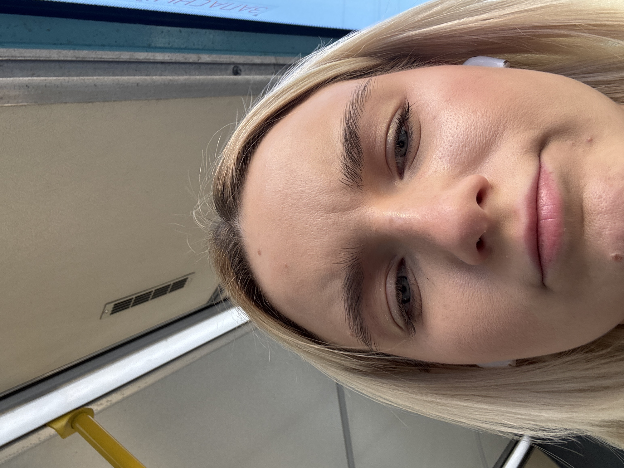 The image shows a close-up selfie of a young woman with blonde hair wearing white earbuds. She appears to be on public transportation, likely a bus or tram, based on the visible interior elements: a window, a railing, and a vent. The location is suggested to be Saint Petersburg, Russia, though there is no direct visual confirmation of this in the image.
