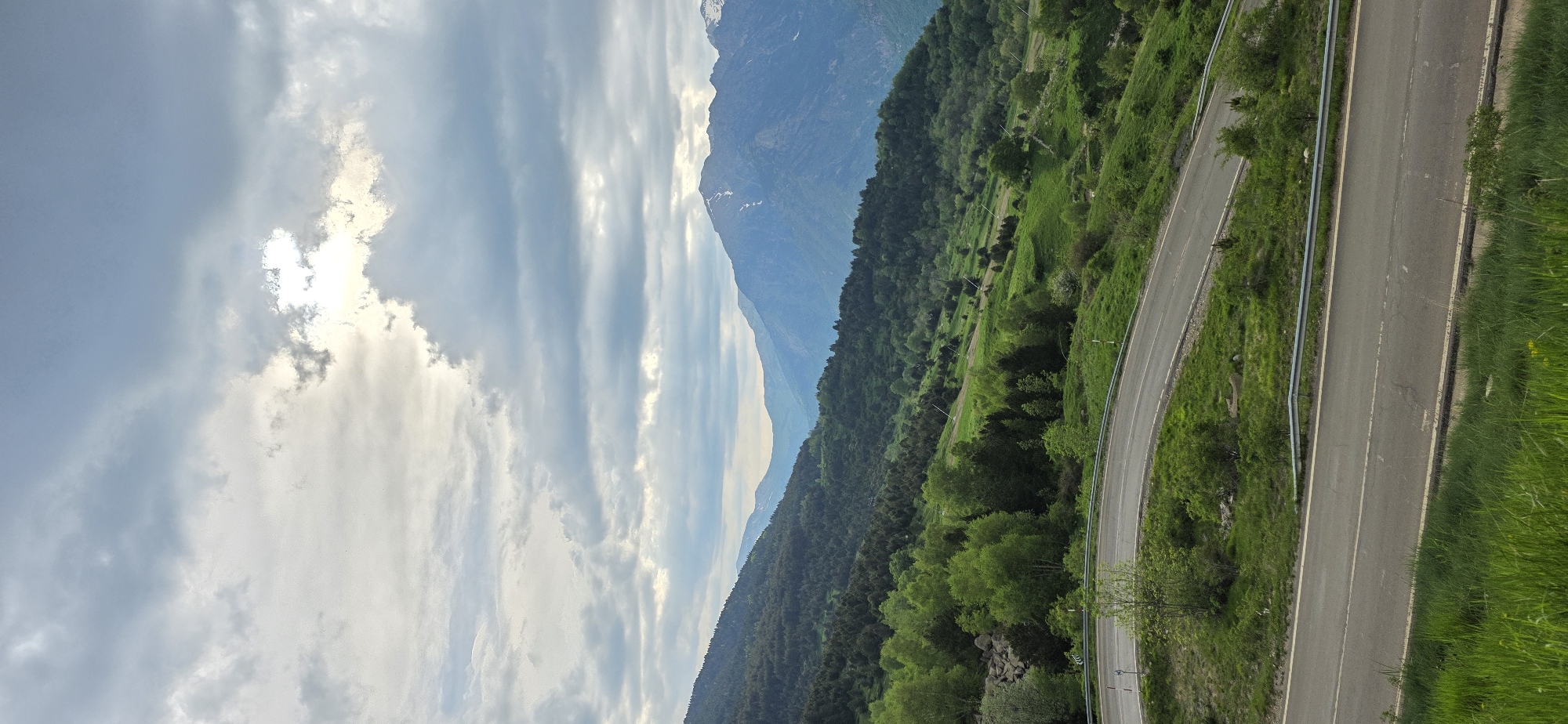 The image presents a landscape near Benasque, Spain, showcasing a mountain range with a visible peak in the background. Cumulus clouds fill the sky. A winding road cuts through a green, mountainous region covered with trees and vegetation. A guardrail runs parallel to the road's edge.