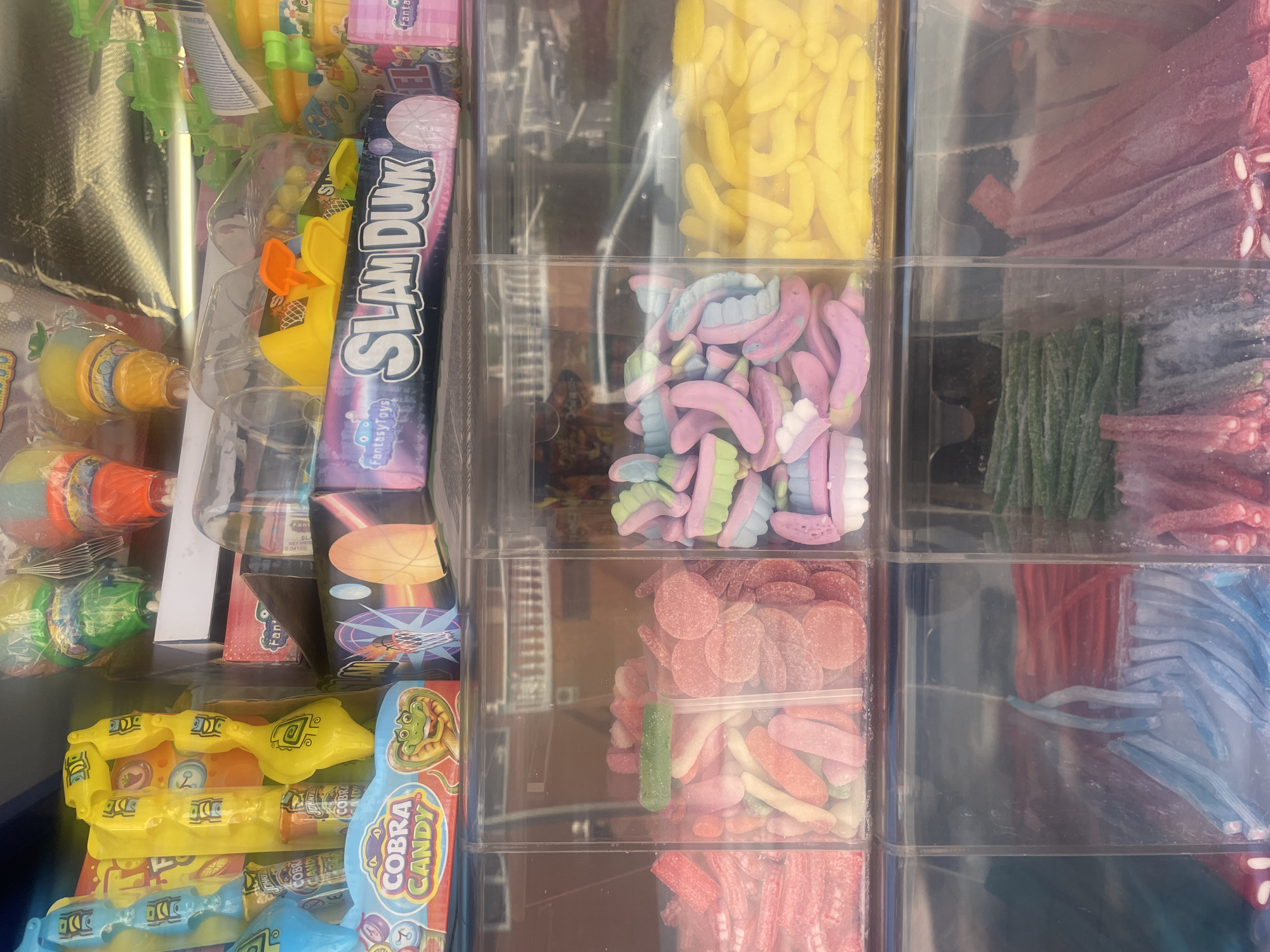 This is a close-up, eye-level photograph of a display of colorful candies and toys for sale, likely at a street vendor or kiosk in Almuñécar, Spain. The display is organized into clear plastic compartments and boxes, showcasing a variety of gummy candies in different shapes and colors, such as red, pink, green, yellow, and multi-colored pastel-toned candies. Some of the candies are in the shape of bananas, circles, and stick-like forms.
Above the gummy candies, there are packaged toys and candies. Prominently displayed are boxes with the text "Cobra Candy" featuring a cartoon snake, and another box labeled "Slam Dunk" with basketball imagery. There are also various individually packaged items, some appearing to be candy lollipops or novelty toys, in bright colors like yellow, green, and orange, some of which are housed in clear plastic packaging. The background is out of focus but suggests an outdoor setting, possibly a street or promenade, with reflections in the glass from the surrounding environment including what appears to be a vehicle. The overall impression is one of a cheerful and vibrant candy selection.