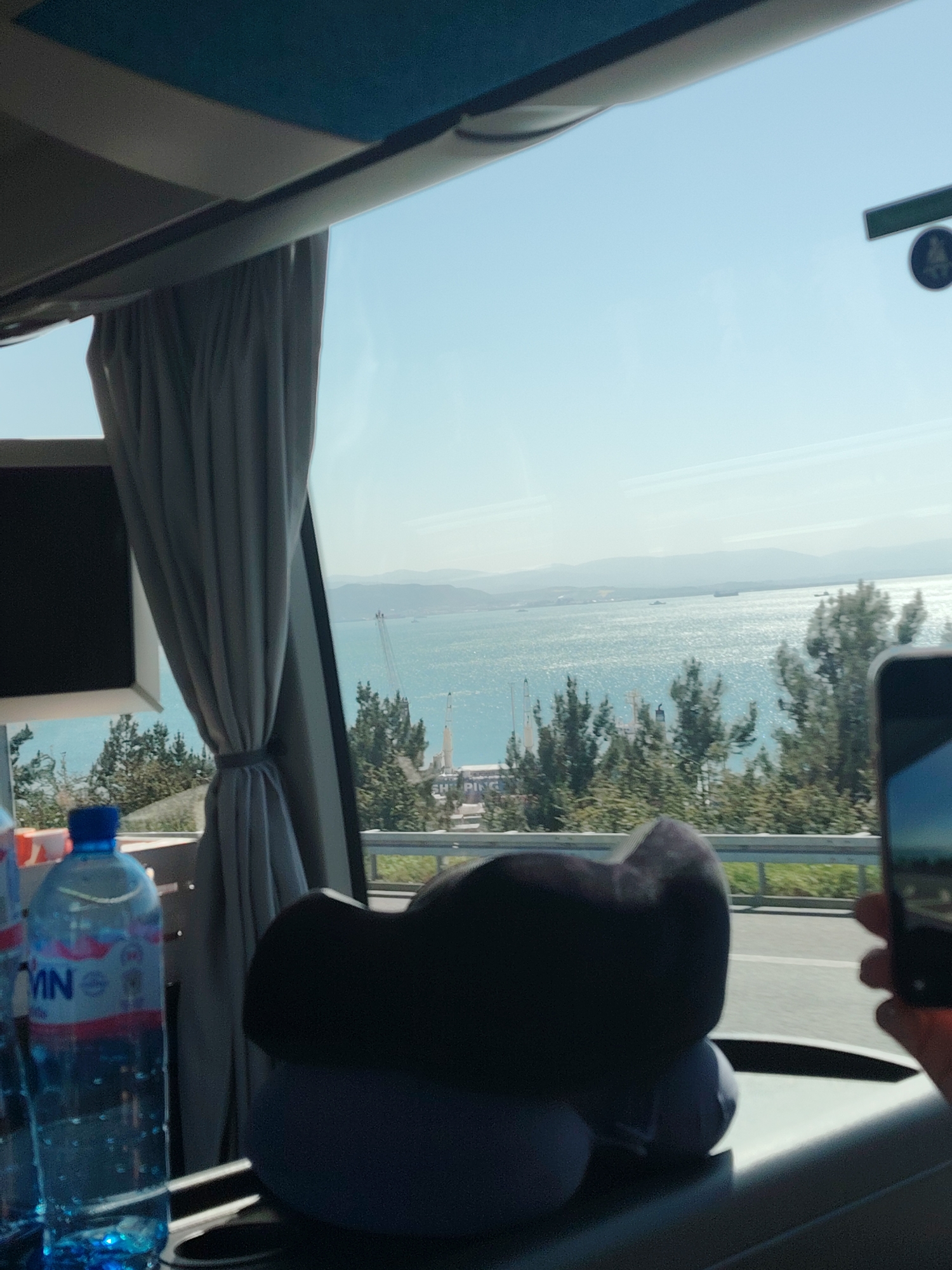 The image captures a bright, sunny daytime scene viewed from inside a vehicle, likely a bus or coach, looking out through a large window. The primary subject is a vast expanse of sparkling blue-green water, consistent with the Marmara Sea in Dilovası, Turkey, stretching towards a distant mountainous or hilly horizon under a clear, light blue sky.
Along the immediate shoreline, green trees are visible, interspersed with industrial structures, including what appear to be cranes and a large white sign partially displaying the word "SHIPPING," indicating a port or industrial area. A road barrier is visible in the lower part of the external view.
Inside the vehicle, a grey curtain is tied back to the left of the window. In the foreground, a dark-colored travel pillow rests on a surface. To the left, a blue plastic water bottle with a blue cap shows the partial text "VIN" on its label, likely part of a brand name like "SİNAN". On the right, a hand holds a smartphone, suggesting the capture of the scenic view. The interior elements of the bus, such as the overhead compartment and window frame, are also visible. A small, circular dark sign with an indistinct symbol (possibly a '4' or an icon) is barely discernible in the upper right. The bright light and reflections on the water suggest it is mid-day or afternoon.