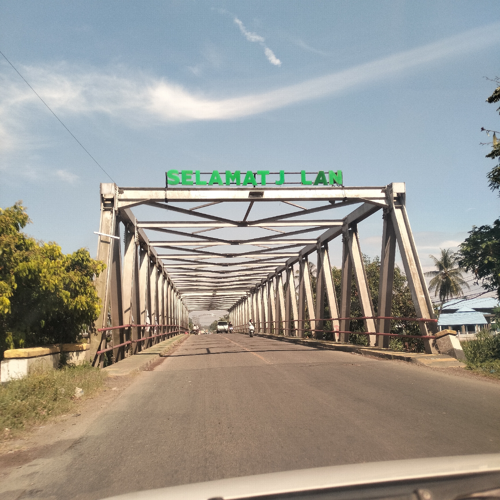 The image presents a daytime view from inside a vehicle, looking straight ahead onto a steel truss bridge in Teupinsiron, Indonesia. The bridge features a sturdy grey metallic structure forming an enclosed passage over an asphalt road. Above the bridge's entrance, prominent green letters spell out "SELAMAT JILAN."
The road surface has faint yellow lane markings, and red metal railings line both sides of the bridge. Traffic is visible in the distance, including a white van and at least two motorcycles. On the left side of the bridge, dense green foliage from trees extends upwards. On the right, more trees, including a palm tree, and a distant building with a distinct blue roof are visible through the bridge's framework.
The sky is clear and bright blue with scattered white cumulus clouds, and a thin power line crosses the upper left quadrant. A faint contrail is also visible. The sunny conditions suggest it is likely late morning or early afternoon. The phrase "Selamat Jalan" translates from Indonesian to "safe journey" or "goodbye," commonly used when someone is departing. The scene depicts routine daily transit across the bridge.