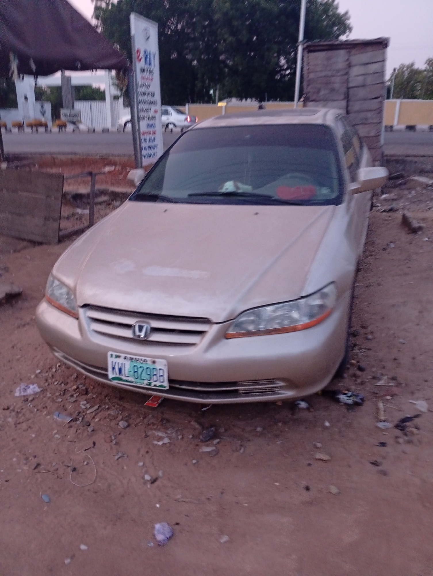 A tan Honda Accord is parked on a dirt lot in Zango-ndaji, Nigeria. The car has a Nigerian license plate, KWL-829BB, and is positioned facing the camera. Debris litters the ground around the car. In the background, there is a street with other vehicles, a sign, and buildings.