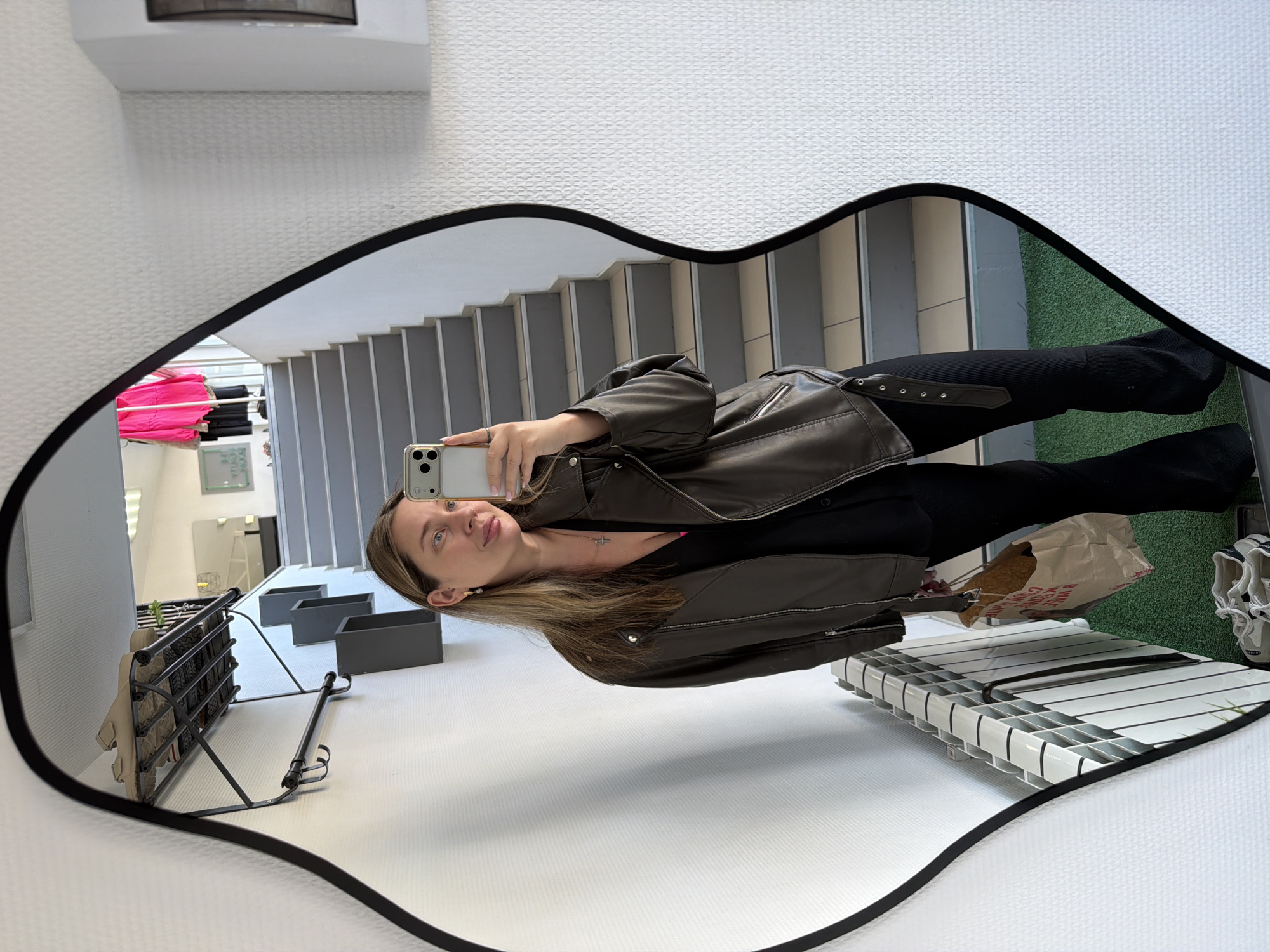 A person is reflected in a lip-shaped mirror while taking a selfie with a smartphone. The person is wearing a brown leather jacket, black top, and black pants. Beside the person is a radiator, a shopping bag, and sneakers on a patch of artificial grass. The mirror also reflects a staircase, shelves, and a white wall with a textured pattern. The location is a building interior, possibly in Chaikovsky, Russia, but this cannot be definitively confirmed from the image.