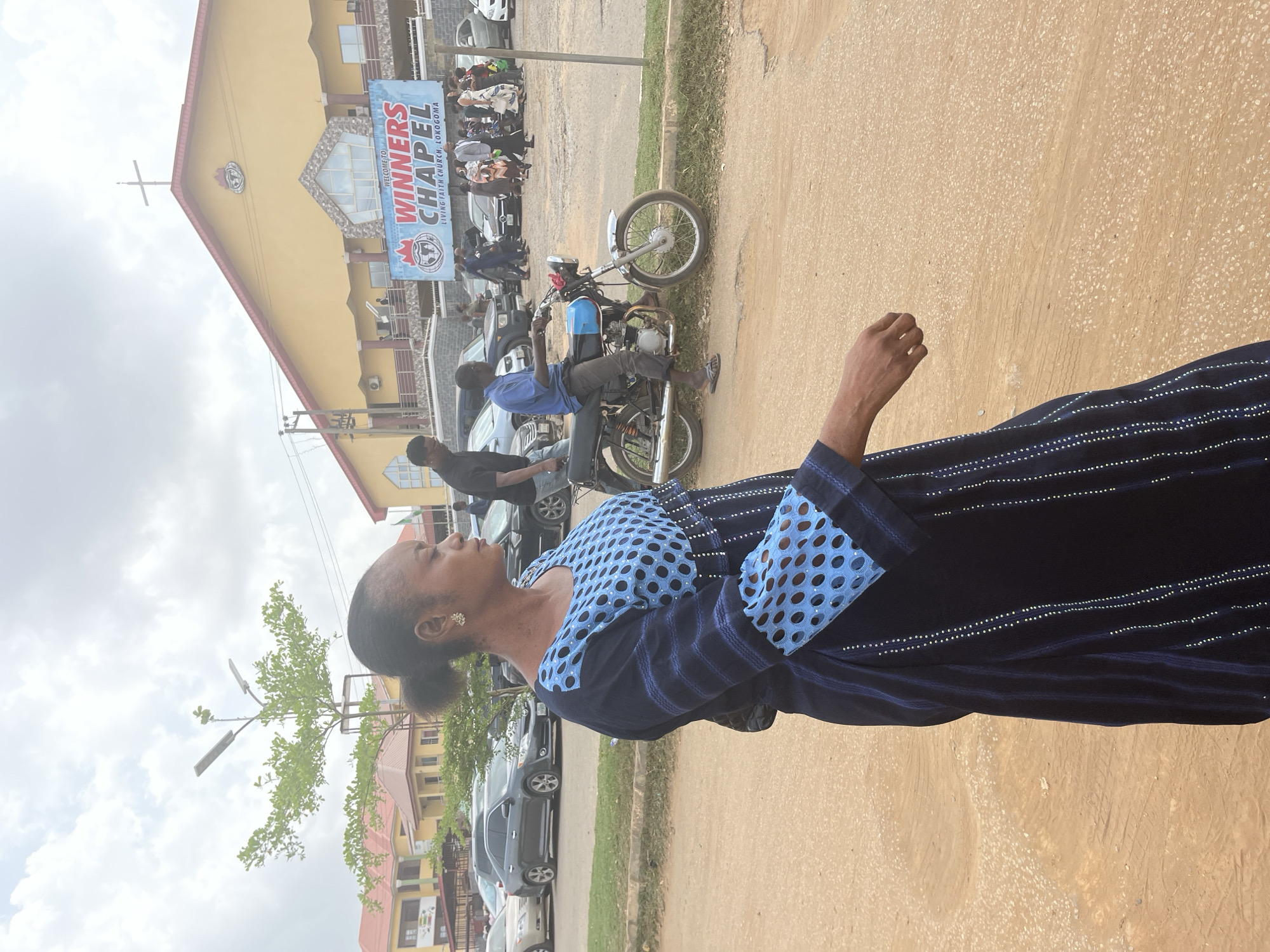 The image shows a woman standing on a paved road in front of Winners Chapel Living Faith Church in Lokogoma, Abuja, Nigeria. She is wearing a navy blue dress with light blue polka dot sleeves and a yoke. A streetlamp and leafy trees are to her left, with parked cars and buildings visible beyond them. In the background, a motorcycle is parked near a crowd of people in front of the church entrance. A banner with "WINNERS CHAPEL LIVING FAITH CHURCH, LOKOGOMA" is visible on the building. The sky is overcast.