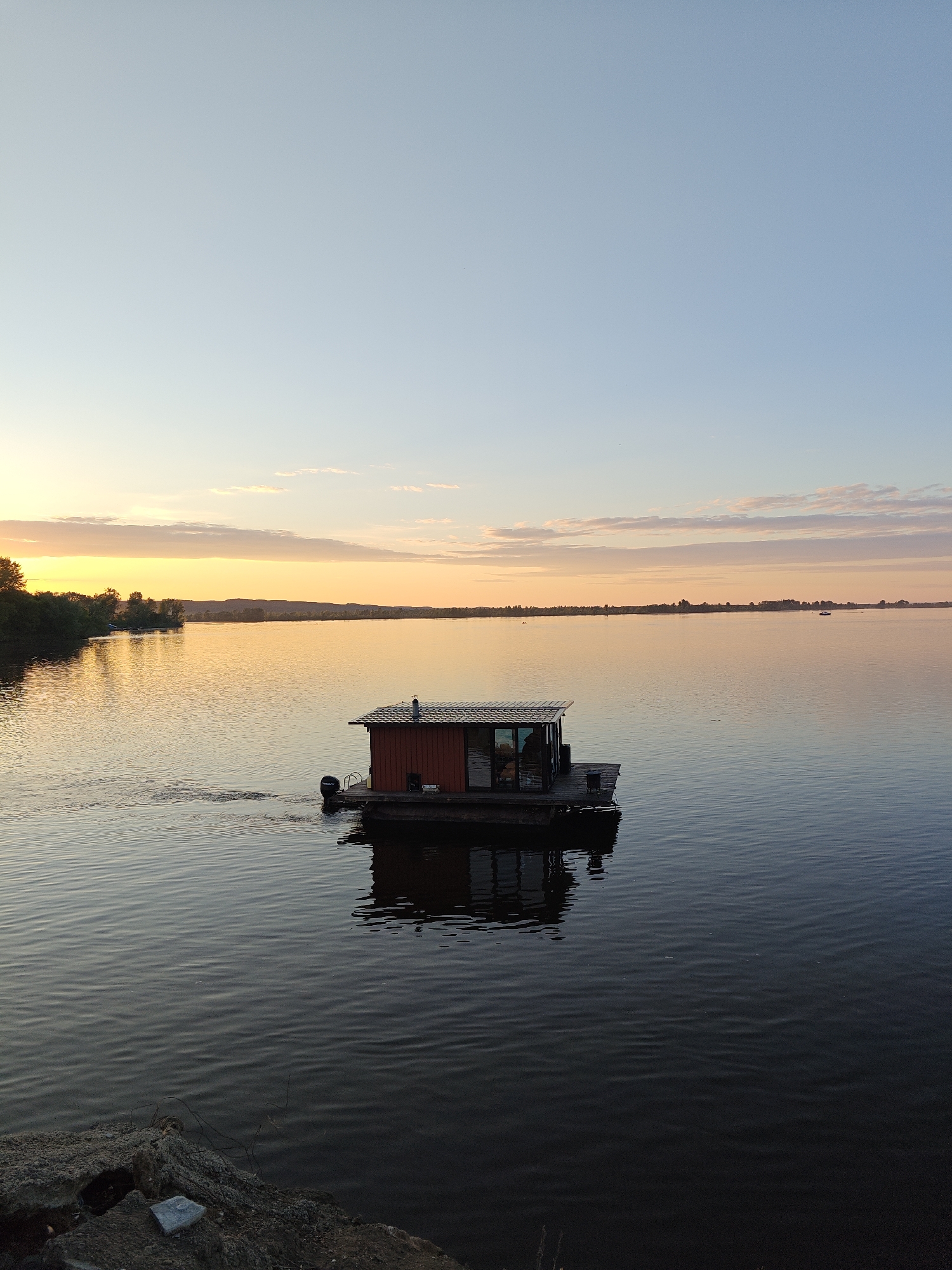 A wide-angle, vertical photograph captures a tranquil scene on a large body of water at either sunrise or sunset, specifically in the area of Nikolo-Beryozovka, Russia. The sky transitions from a pale blue at the zenith to a warm golden-orange and pink closer to the horizon, dotted with sparse, elongated clouds.
The central subject is a small, rectangular house-like structure, painted reddish-brown, situated on a dark floating pontoon or raft in the middle distance. This structure features a dark, shingled roof with a prominent silver chimney pipe. Large glass windows or doors are visible on the side of the building, reflecting the ambient light. A black Mercury brand outboard motor is attached to the stern of the pontoon, generating a discernible wake on the water's surface, indicating recent movement or current propulsion.
The water reflects the sky's changing colors, appearing golden-orange towards the left horizon and darker blue-grey in the foreground and to the right. Ripples cover the entire surface. To the left, a dense, dark green treeline marks the nearest bank, silhouetted against the glowing sky. Along the distant horizon, low hills and scattered trees are faintly visible, and at least one other small, dark boat can be discerned on the water in the far right distance.
The immediate foreground, at the bottom left, reveals a rough, earthy bank composed of dark, uneven rock or concrete debris, with a small, light-colored stone resting on it. No individuals are visible in the image.