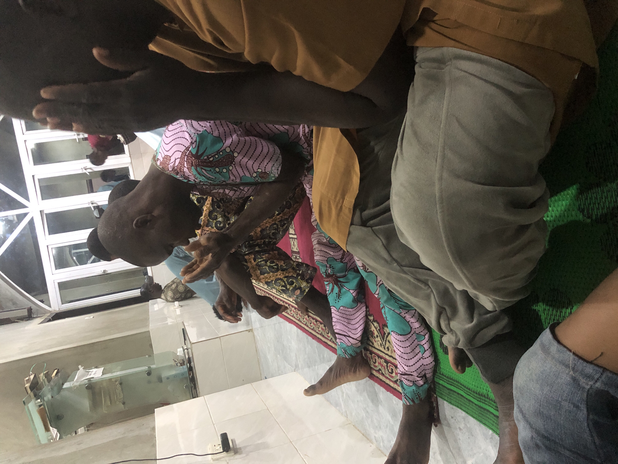 In Ado, Nigeria, a group of men are engaged in prayer. The scene is indoors, likely a prayer hall or mosque, with white tiled walls. Several men are seated on prayer mats, which are layered on the floor. One man, in the foreground and slightly to the left, is wearing light grey sweatpants and is positioned on a green mat. Another man, to his right, wears brightly patterned pants with pink and teal designs, and is seated on a red rug. A third man, in the far right of the image, is wearing a tan-colored garment. The men appear to be in a state of prayer, with hands raised in supplication in the case of the man in the patterned pants. In the background, a glass structure, possibly a lectern or display case, is visible against the wall. Large windows with white frames are also present in the background, suggesting the presence of natural light or an exterior view. The lighting in the room is artificial, casting a warm glow.