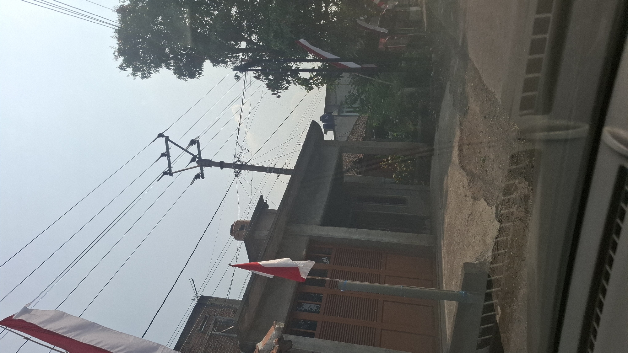 The image shows a street scene in Warungkondang, Indonesia. In the foreground, a cracked and uneven asphalt road is visible, with a concrete barrier on the left. The middle ground features a building with a brown wooden garage door and a covered entrance. Two Indonesian flags, red and white, are prominently displayed on poles in front of the building and further down the street. Overhead, electrical wires stretch across the sky, supported by a utility pole. The background reveals more buildings, lush green trees, and a hazy sky. The overall impression is of a residential area on a sunny day.