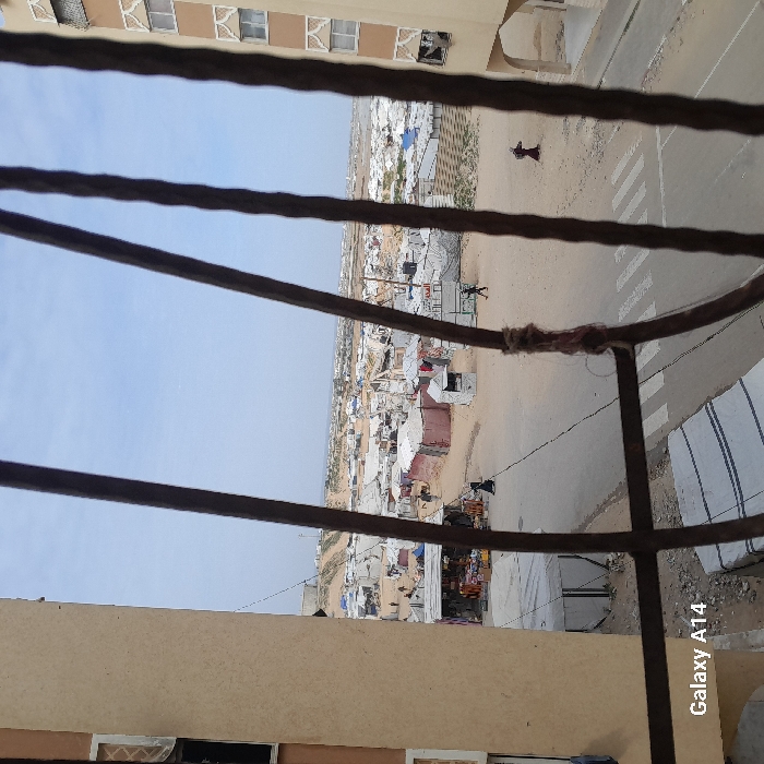 The image depicts an expansive informal settlement in Khan Yunis, Palestinian Territory, viewed from an elevated position, looking out through four dark, vertically oriented metal window bars.
In the immediate foreground, a portion of a light-tan building wall is visible on the left, and another section of a similar building with decorative white window frames and arches is on the right. Dark metal bars, one wrapped with a piece of brown twine, bisect the frame vertically. The bottom left corner displays a "Galaxy A14" watermark.
The midground features a wide, unpaved dirt road with faded white crosswalk markings. Along this road and extending into the background, a sprawling settlement of numerous makeshift shelters is visible. These structures are predominantly constructed from white or light-colored tarpaulins and sheeting, with some incorporating reddish-brown materials. Several small, open-front shops or stalls line the road, displaying various goods.
People are observed moving within the scene. On the dirt road, one individual in a dark abaya walks from left to right near a shop. Another person in dark clothing walks left to right further down the road, while a third individual, also in dark attire, walks from right to left on the opposite side of the road. Other figures are faintly visible deeper within the camp.
The background consists of more makeshift shelters extending across undulating sandy terrain and low hills, merging with a pale blue, hazy sky. Distant structures or elements of a more permanent settlement are vaguely discernible on the horizon. The environment appears arid and densely populated with temporary housing structures.