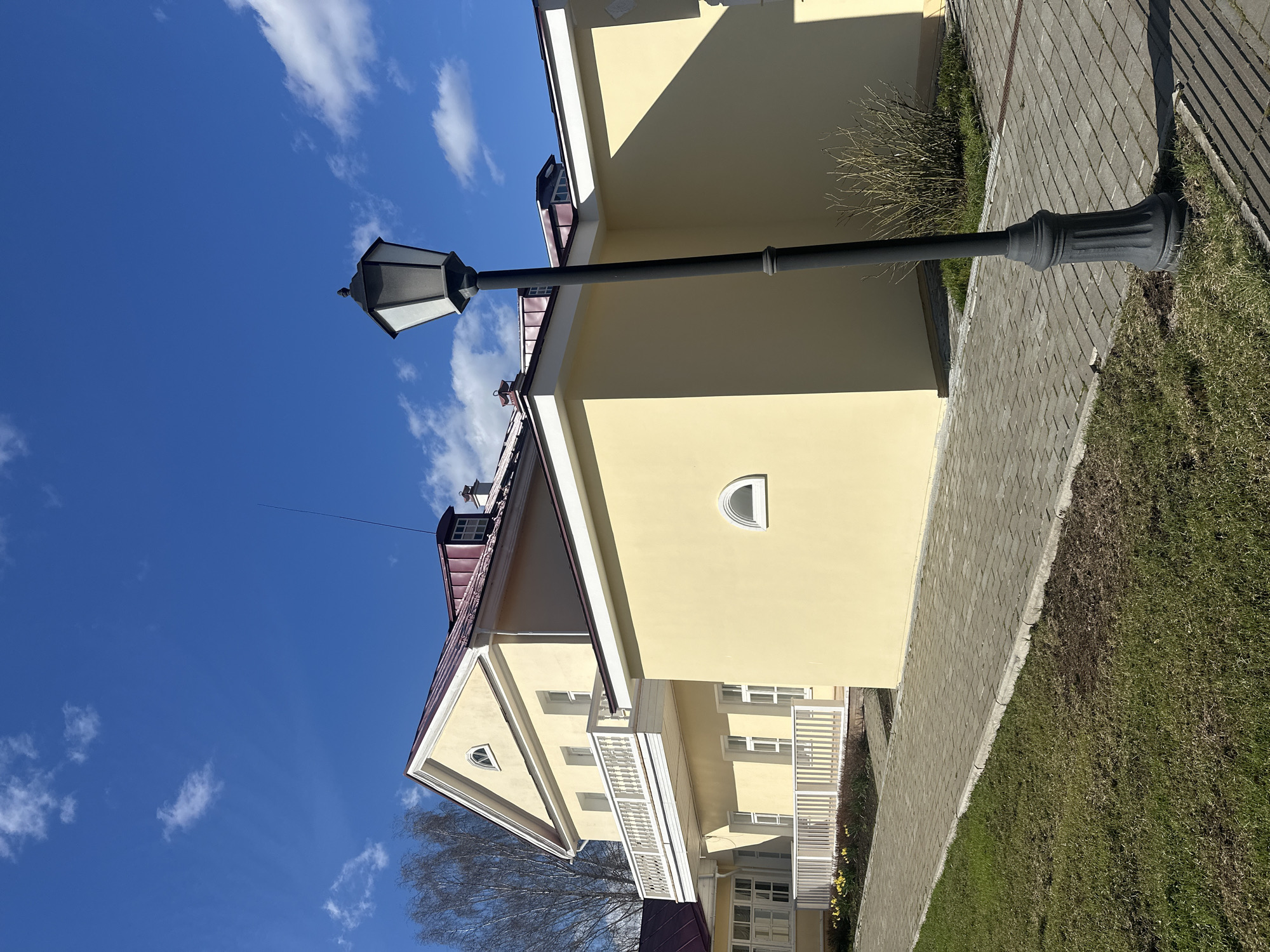 The image shows a cream-colored two-story building in Votkinsk, Russia, under a bright blue sky with scattered white clouds. The building has a maroon roof, white trim, and a white balcony. A black street lamp stands in front of the building, its base situated on a paved walkway bordered by grass. The photograph is taken at an angle, with the building appearing diagonally.
