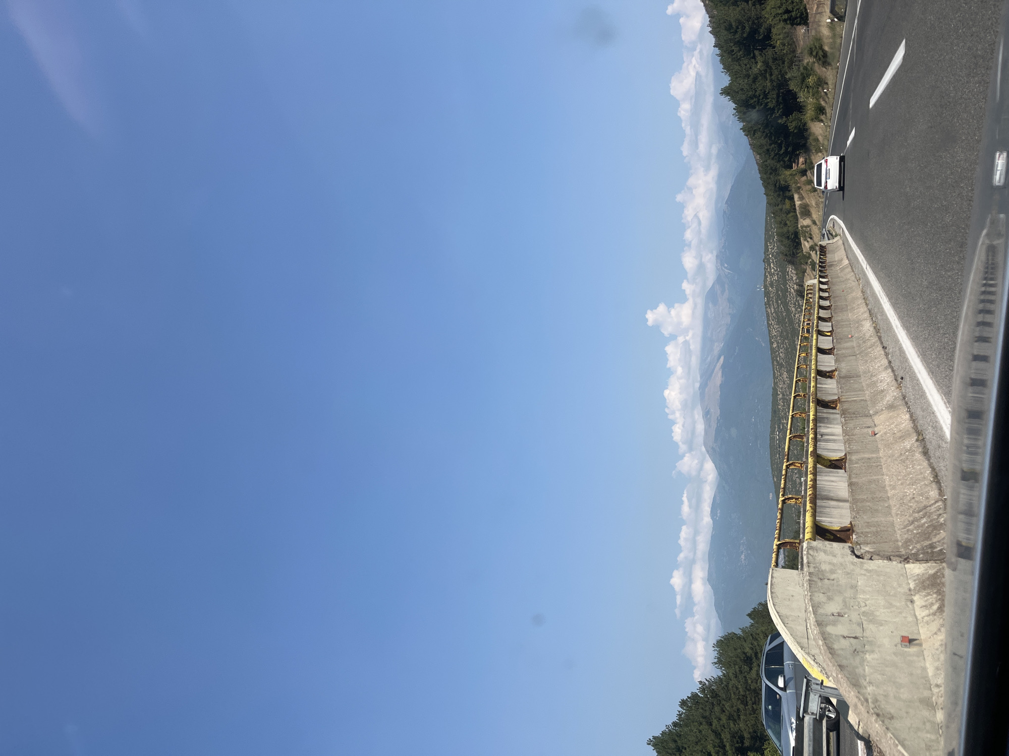The image depicts a view from inside a vehicle traveling on a highway in Zhelino, North Macedonia, on a clear, sunny day. The highway has two lanes of asphalt with white lane markings. A concrete barrier with a rusty yellow guardrail separates the lanes. On the left side of the highway, a silver SUV is parked or stopped. Beyond the barrier, a dense forest of evergreen trees lines a hillside. In the distance, a range of majestic mountains under a bright blue sky with scattered white clouds is visible. The lower part of the image shows the dashboard and windshield reflection of the vehicle from which the photo was taken.