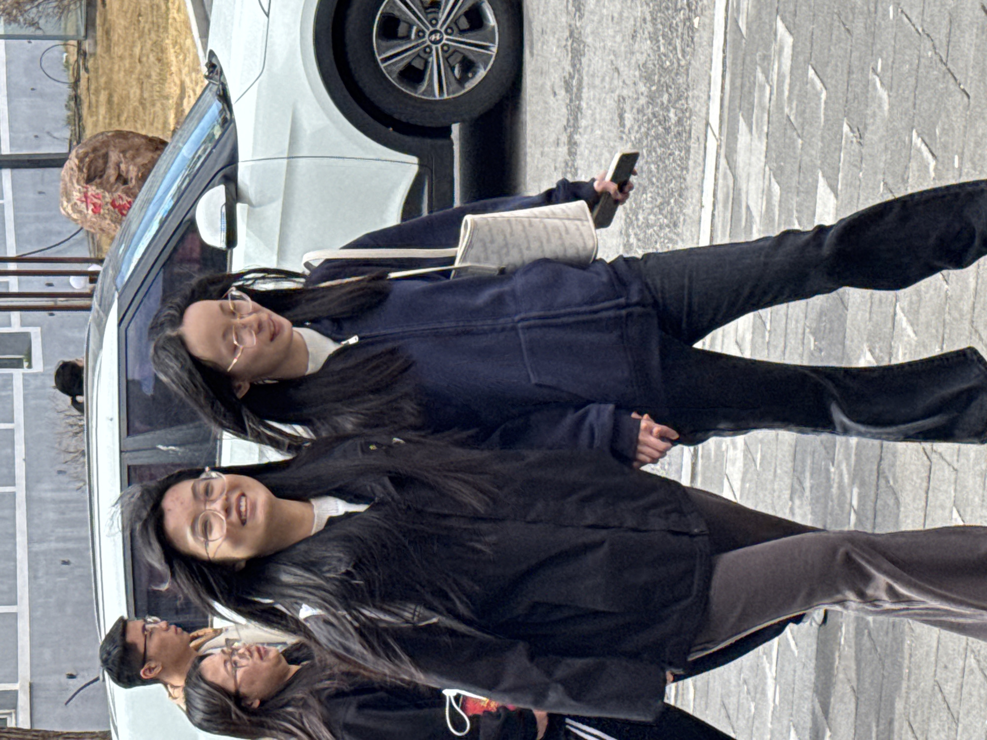 Two young women are walking on a sidewalk paved with grey bricks. The woman in the foreground is wearing a black jacket and grey pants, while the woman behind her has on a blue jacket and black pants. Both are wearing eyeglasses and have long dark hair. The second woman is holding a smartphone. To their left is a white car parked alongside the sidewalk. The background includes some greenery and a building. The location appears to be Zhongtun, China, based on the context.