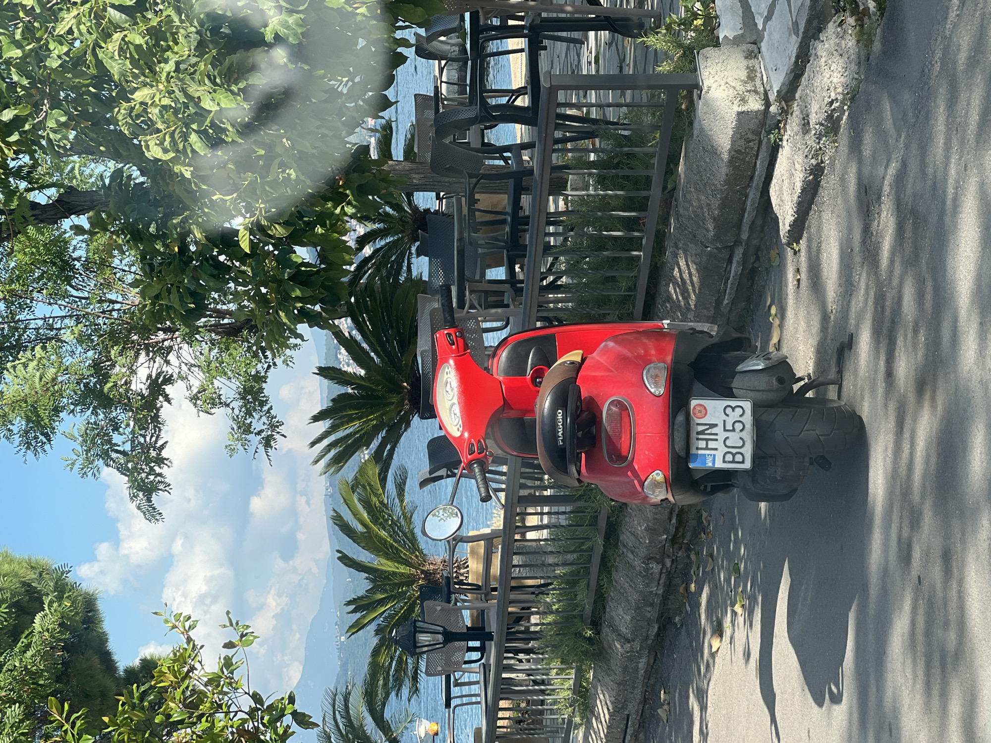 This image, taken in Đenovići, Montenegro, captures a sunny outdoor scene featuring a parked red scooter and a waterfront area.
In the foreground, a vibrant red Piaggio scooter is parked on a paved road or sidewalk, casting a distinct shadow to its right. The scooter's license plate, "MNE HN BC53," is clearly visible on its rear. To the left of the scooter, a metal railing separates the road from a terraced area with outdoor seating. Several tables and chairs, some stacked, are arranged behind this railing, suggesting an outdoor cafe or restaurant. Lush green foliage, including palm trees and other deciduous trees, frames the scene, with some branches extending overhead and casting dappled shadows.
Beyond the railing and cafe area, a calm body of water—likely the Adriatic Sea, given the location context—stretches into the distance under a bright blue sky dotted with fluffy white clouds. Mountains rise in the background across the water, adding depth to the scenic vista. The strong sunlight and clear sky indicate a pleasant day, likely midday or early afternoon, with distinct shadows visible on the ground from the scooter and trees. No people are visible in the frame, creating a serene, inviting atmosphere.