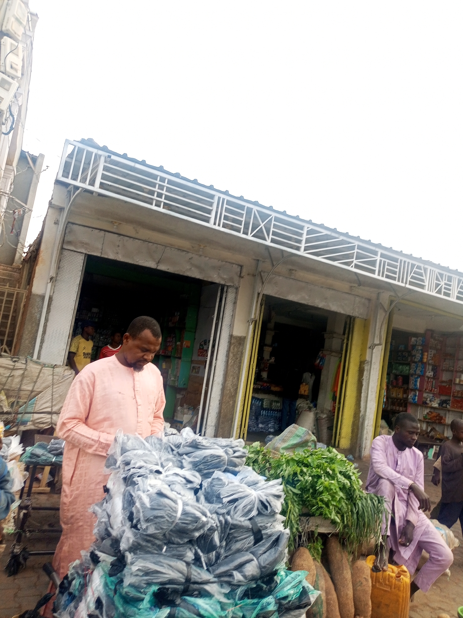The image depicts a street scene in Tempure, Nigeria, with a row of small storefronts visible in the background. In the foreground, an African man wearing a peach-colored robe is standing next to a large pile of items, seemingly bags wrapped in plastic, possibly for sale. Adjacent to him, another man in a lavender robe is seated beside a display of fresh vegetables and what appears to be yams. A yellow jerrycan sits nearby. A child is partially visible in the background. The stores in the background appear to be stocked with various goods. The sky is overcast.
