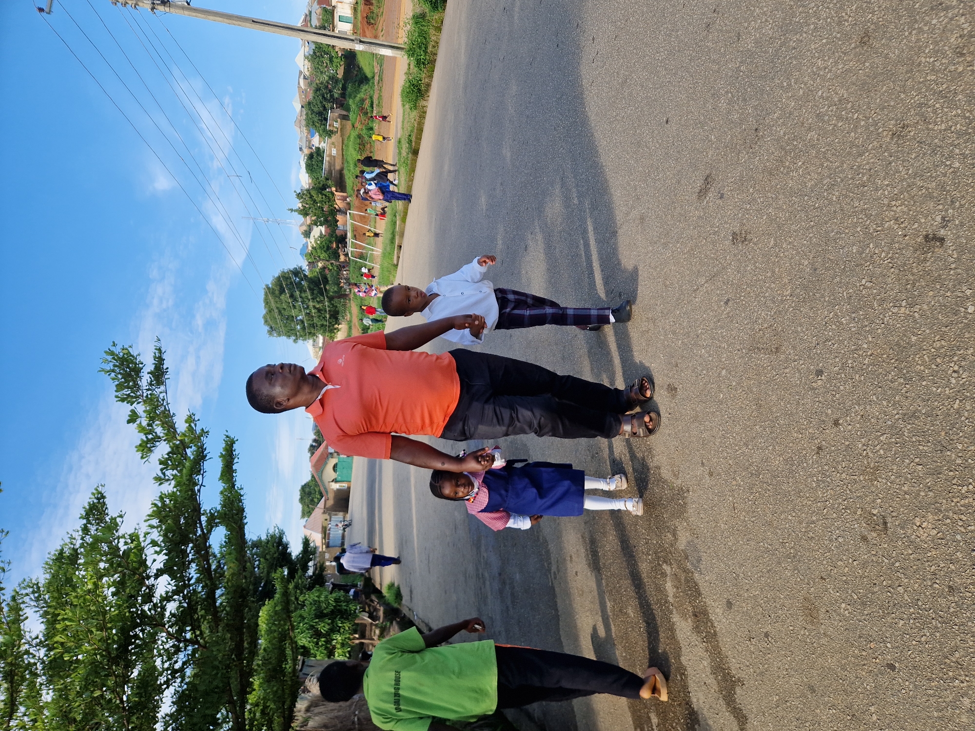 A man in an orange polo shirt and black pants walks hand-in-hand with a young girl wearing a blue dress with white trim and a young boy in a white collared shirt and plaid pants, on a paved street. The man has dark skin and is wearing sandals. The girl is holding the man's left hand, and the boy is holding the man's right hand. To the left, a person in a green t-shirt and black pants walks away from the camera. In the background, there are houses with corrugated metal roofs, trees, and utility poles. Further in the distance, a group of people are gathered around a small soccer field. The scene is set in Kabong, Nigeria, under a bright blue sky with scattered clouds.