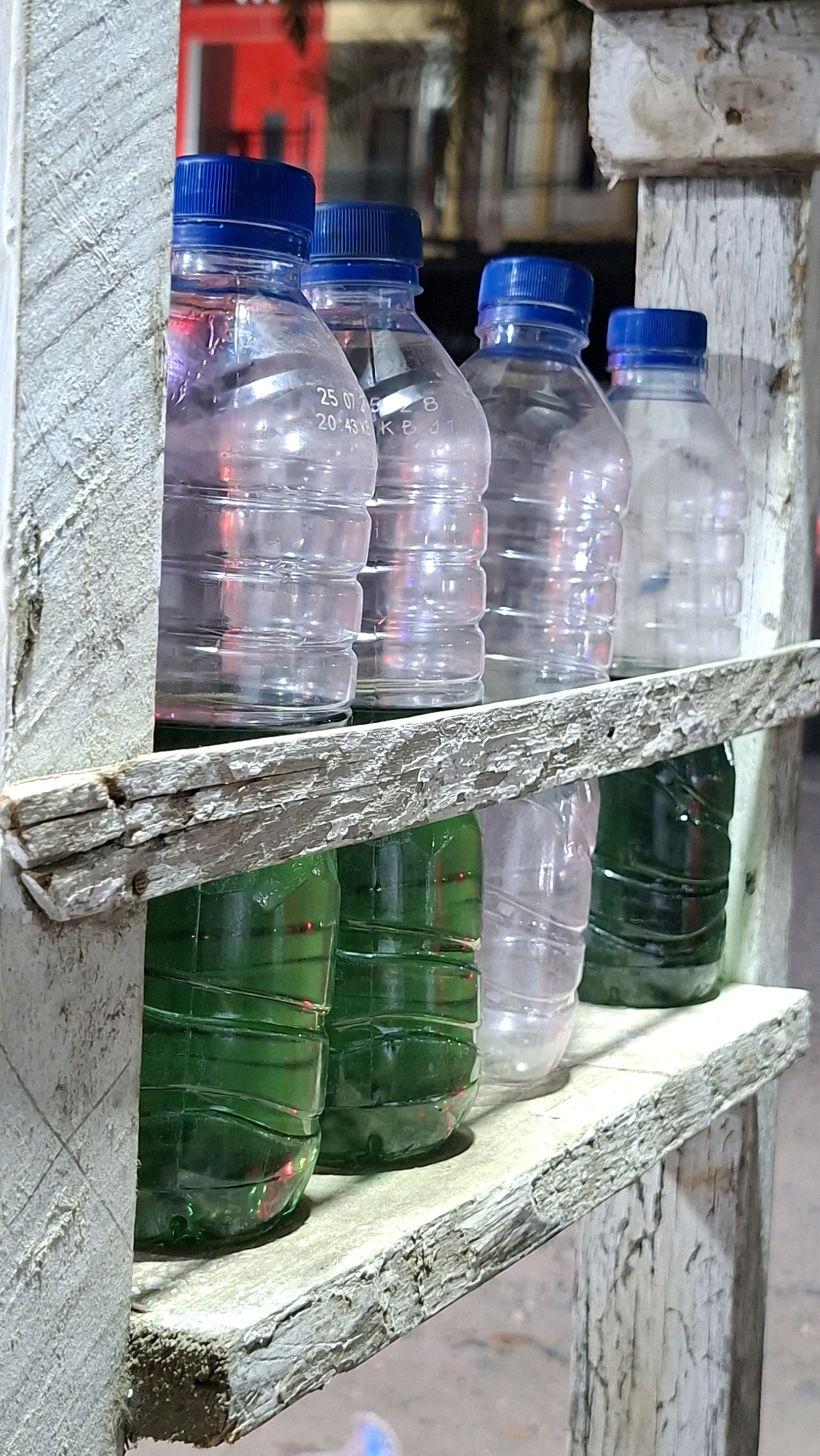 The image shows a close-up of a wooden shelf structure displaying several plastic bottles. The bottles are arranged on two shelves. The upper shelf holds five clear plastic bottles with blue caps, some of which contain a clear liquid. Faint text is visible on one of these bottles. The lower shelf holds at least three green plastic bottles, also with blue caps. The wooden structure appears weathered and aged. The background is blurred, but suggests an outdoor urban environment with hints of red buildings and greenery, possibly palm trees. The lighting is somewhat dim, with some red and yellow reflections on the bottles. The scene is located in Lhokseumawe, Indonesia.