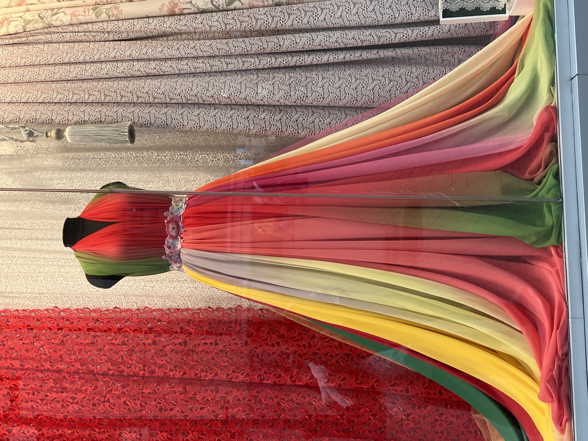A mannequin is displayed in a shop window, wearing a flowing, multi-colored chiffon dress. The dress transitions through various shades, starting with green and red at the top, then a gradient of red, pink, orange, yellow, and light green towards the hem. The mannequin is adorned with a decorative belt. Behind the mannequin, a backdrop of textured fabric curtains can be seen. To the left, a bright red lace curtain is visible. To the right, there are neutral-toned curtains with a floral pattern, and a light blue tassel hangs in the foreground. The scene appears to be a retail display in a store.