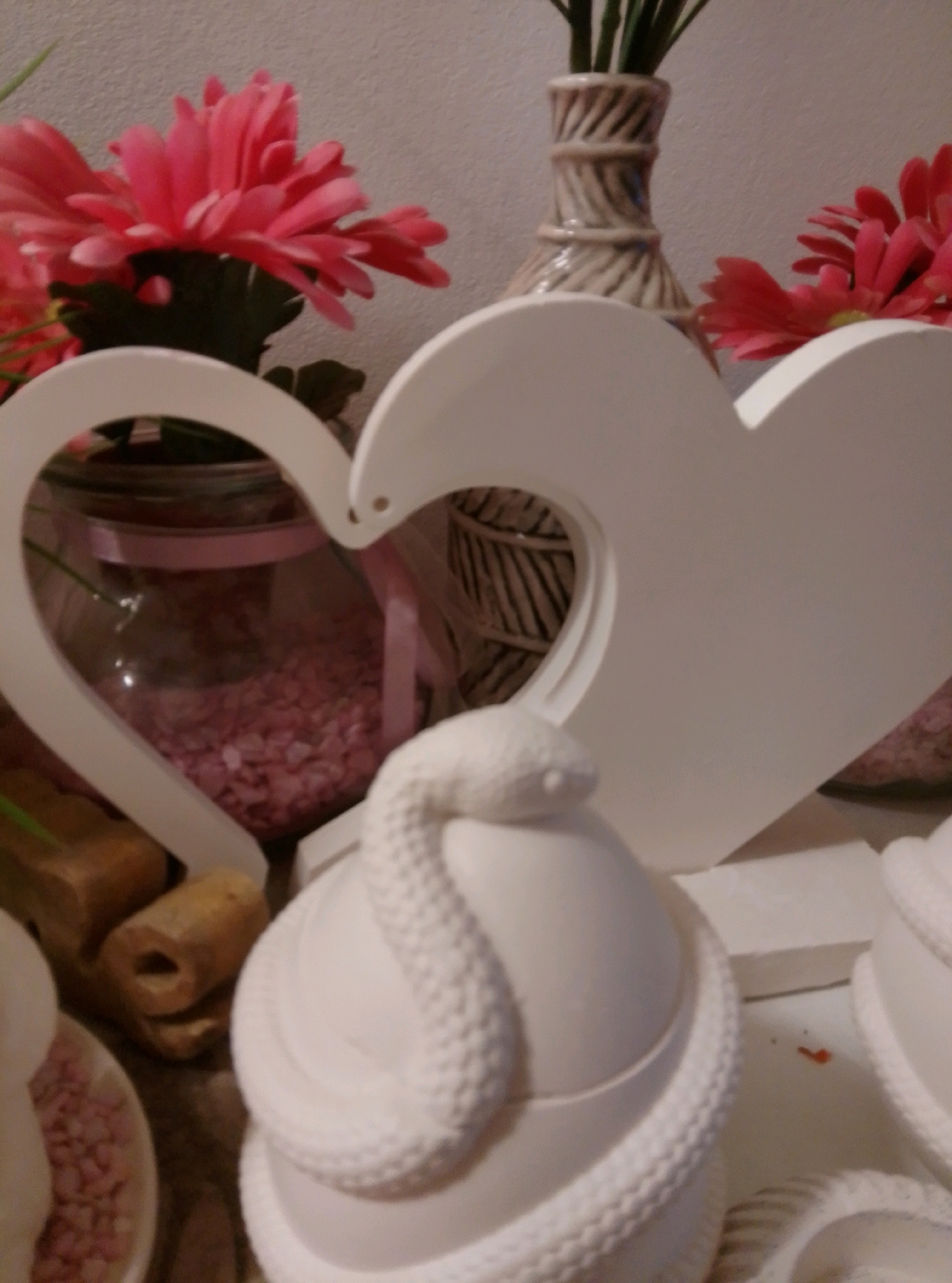 This is a close-up, eye-level, indoor shot of a white ceramic decorative heart cutout framing a glass jar filled with pink potpourri and ribbon. To the left of the jar, a pink artificial flower is visible, and behind the jar, a textured brown vase with greenery is present. In the foreground, a white ceramic snake coiled around an egg-shaped object is partially visible. There are no people in the image, and the setting does not provide enough information to determine a specific location within the city of Wedel, Germany.