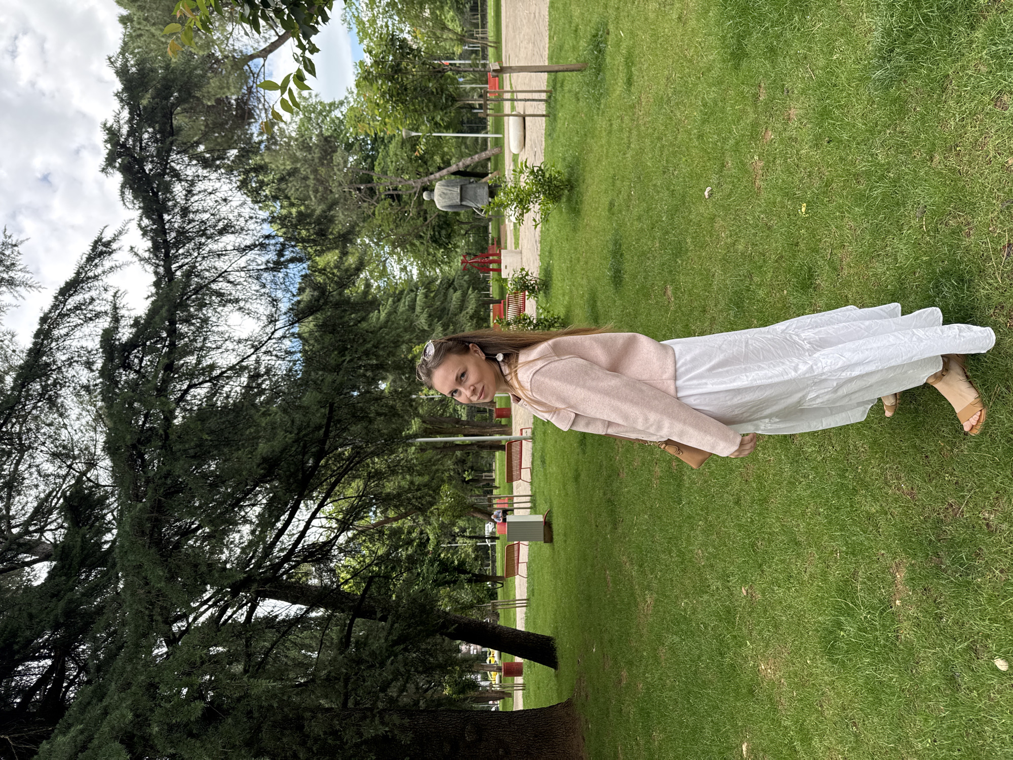 The photograph shows a woman standing on grass in a park-like setting. She is wearing a white dress and a light pink jacket. Trees are visible in the background. Benches can be seen amongst the trees in the background. A statue is also visible in the background. Based on the environment, this is likely in a park in the city of Podgorica, Montenegro.