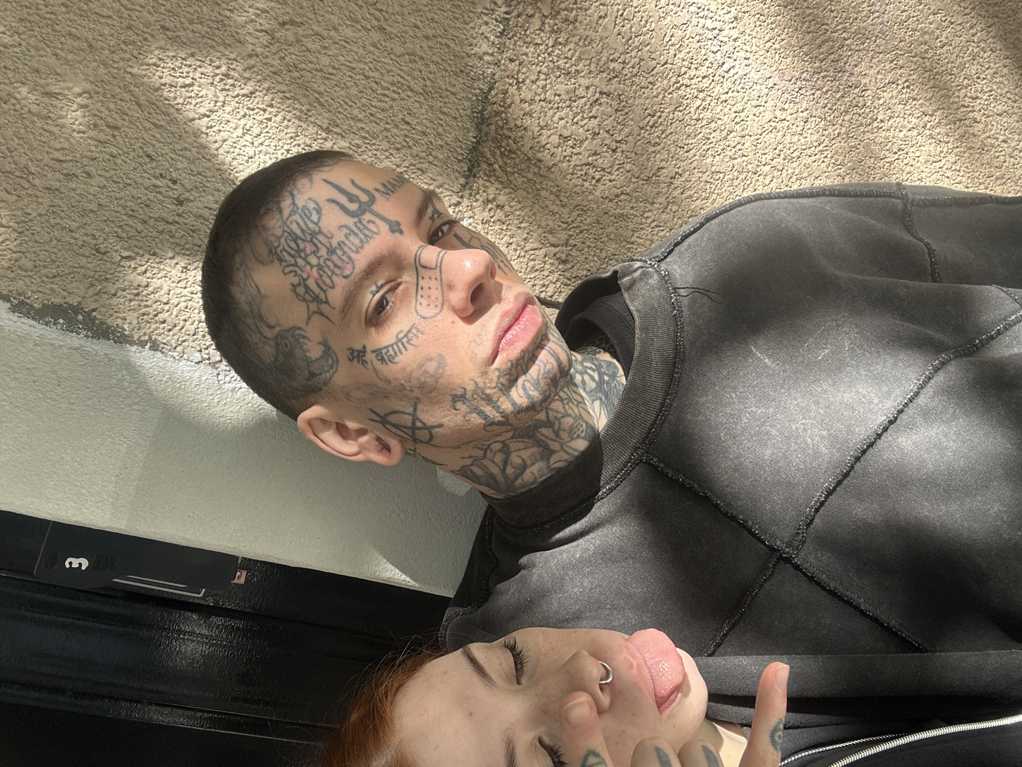This close-up, high-angle shot features two individuals, a man and a woman, seemingly lying down or reclining in bright daytime sunlight.
The man, positioned centrally in the upper half of the frame, has short dark hair and extensive black tattoos covering his face, head, and neck. Notable facial tattoos include a trident symbol on his forehead, elaborate script resembling "Love" on his temple, a drawn band-aid design on his nose/cheek, and Sanskrit (Devanagari) script beneath both eyes. Specifically, "अहं ब्रह्मास्मि" (Aham Brahmasmi, meaning "I am Brahman") is visible below his right eye, and "महाकाल" (Mahakal) below his left eye. His neck features intricate floral or rose-like tattoos. He wears a dark grey, distressed-looking sweatshirt with prominent visible stitching, and gazes directly at the camera with a neutral expression.
Partially visible in the lower portion of the frame is a woman with red hair. She wears a silver nose ring and is captured with her tongue playfully sticking out. Her visible clothing is dark, possibly black, with a zipper detail. Her fingers, which are also tattooed, are partly shown near her mouth.
The background above the man consists of a light-colored, textured wall or surface, possibly stucco or concrete, strongly illuminated by sunlight casting distinct shadows. Below the subjects, there is a dark, glossy, horizontal surface, potentially a railing or piece of furniture. A black card or tag with the number "3" on it is tucked near the man's ear, against this dark surface. The image conveys an intimate and somewhat playful mood. While the provided context suggests Buenos Aires, Argentina, there are no explicit visual cues within the image to confirm this location.