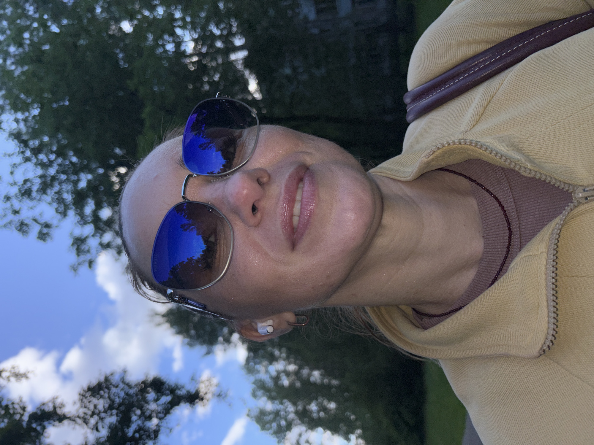 A medium shot captures a woman from the chest up, outdoors, possibly in a park in Sinyavino, Russia, given the surrounding greenery and sky. She is wearing large, reflective sunglasses that mirror the blue sky and trees. Her head is tilted slightly upwards and to the right. She has on a light yellow zippered jacket over a mauve-colored long-sleeved shirt. A thin, dark beaded necklace is visible around her neck. A dark brown strap, possibly from a bag, rests on her right shoulder. In her left ear, a white wireless earbud is visible. The background consists of blurred green foliage and a bright blue sky with scattered white clouds. The lighting suggests it is daytime, with the sun reflecting strongly off her sunglasses.