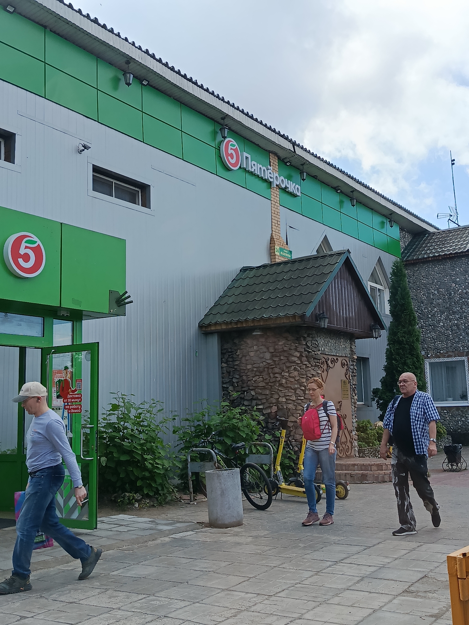 The image depicts the exterior of a "Pyaterochka" supermarket in Orekhovo-Zuyevo, Russia, under a cloudy sky. The building features a prominent green facade with the red and white "5" logo. Three individuals are visible: a man wearing a cap and blue jeans is walking towards the entrance on the left, carrying a shopping bag; a woman with a red backpack and striped shirt is walking in the middle; and a man in a blue checkered shirt and patterned pants is walking on the right. In the background, there are two yellow electric scooters and a bicycle parked near the entrance, along with some greenery and stone-clad architectural elements. The ground is paved with interlocking concrete tiles.