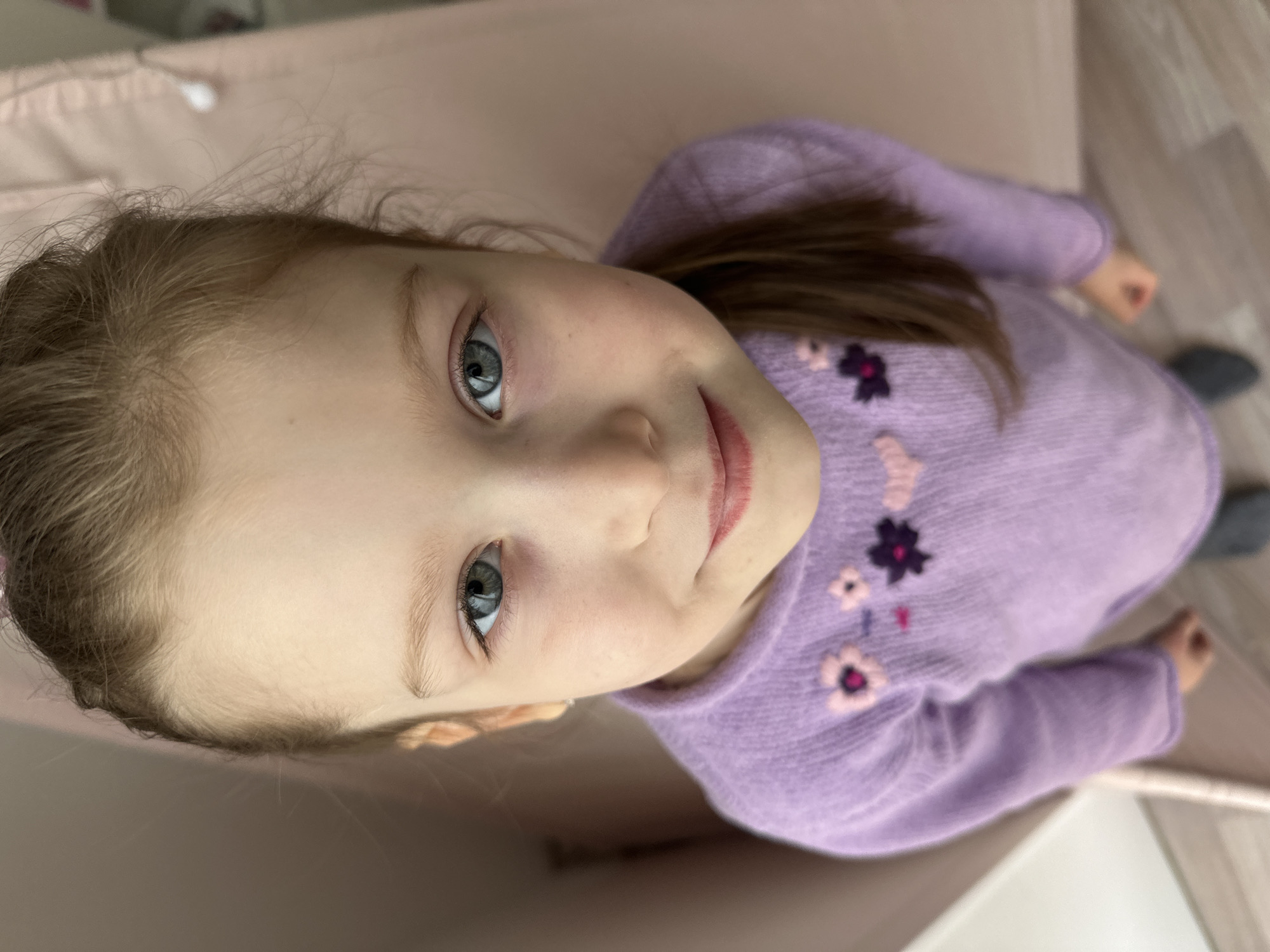 A young child with fair skin, light blonde hair, and light blue eyes wearing a light purple long-sleeve sweater with pink and dark purple floral designs is looking up at the camera. The background shows a light pink or beige surface and part of a light-colored floor. The location is unknown and there is no information available to determine if it's Chaikovsky, Russia.