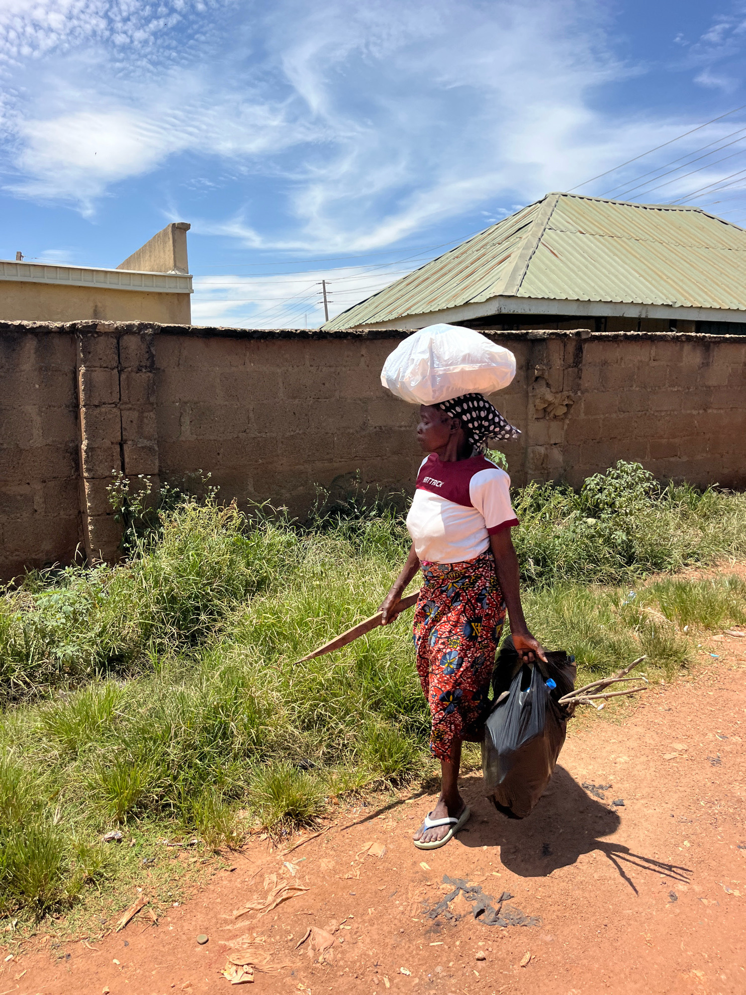 A woman walks on a dirt road in Jos, Nigeria, carrying a large white bag on her head, a wooden stick in her right hand, and two black plastic bags filled with sticks in her left hand. She is wearing a white and maroon t-shirt, a colorful patterned skirt, and flip-flops. The scene is outdoors, with a brick wall and overgrown vegetation in the background. The sky is blue with scattered clouds.