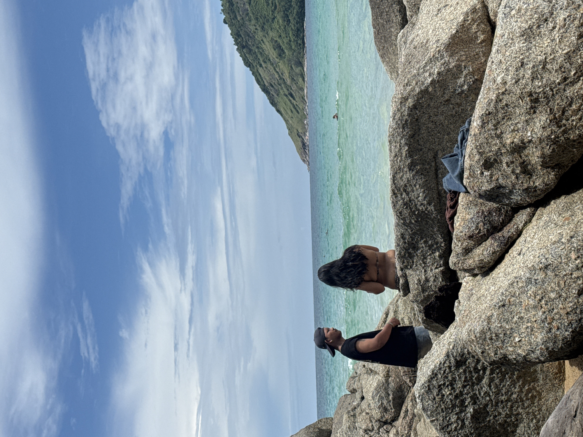 Two individuals are situated on a rocky shoreline overlooking a turquoise ocean under a partly cloudy blue sky. On the left, a person wearing a black t-shirt and a grey baseball cap faces towards the right, appearing to be in conversation. To their right, a person with long, dark hair is seated, wearing a patterned bikini top and facing away from the camera, looking out at the water. In the distance, a green, tree-covered hill slopes down to the sea. The water is clear and shows small waves. In the mid-ground of the ocean, a lone swimmer is visible. The foreground is dominated by large, rough-textured granite boulders. A dark brown cloth and a blue garment are draped over one of the boulders in the foreground. The overall setting is a scenic coastal area, likely within Rawai, Thailand, known for its rocky beaches and picturesque views.