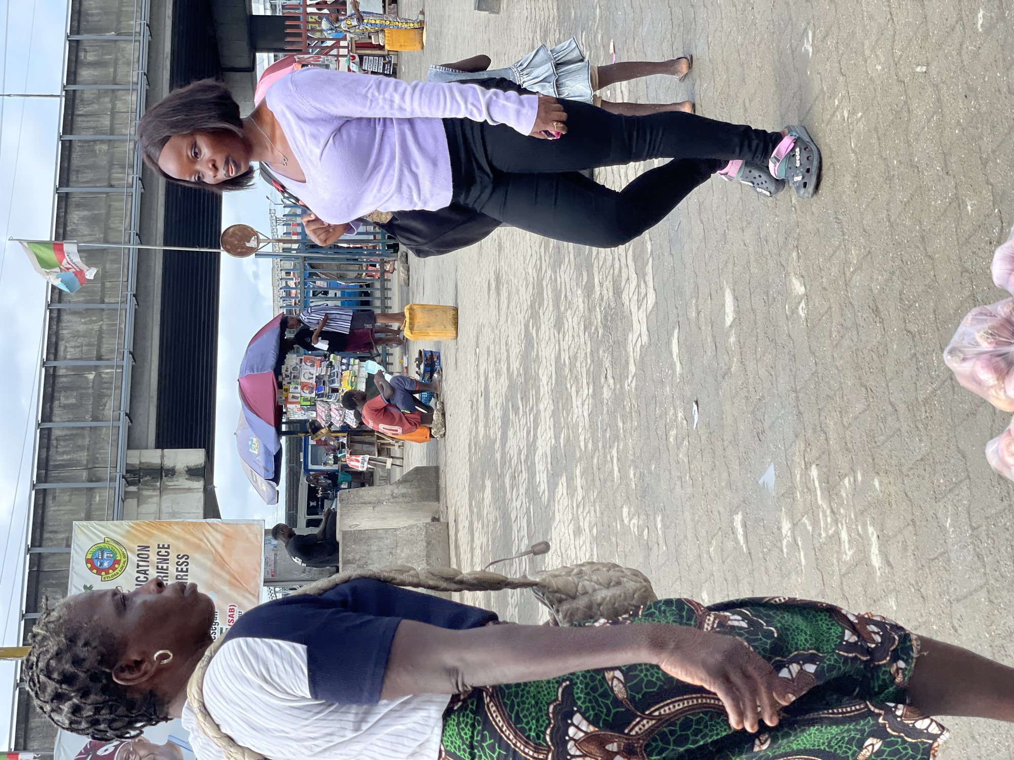 The image depicts a street scene in Lekki, Nigeria. In the foreground, an elderly woman in a patterned green and brown skirt, a blue shirt, and a white strap across her shoulder is visible. A younger woman wearing a lilac long-sleeve shirt and black leggings is standing behind her, facing the camera. The ground is paved with rectangular blocks. Further in the background, there are vendors with umbrellas and various goods for sale. A building structure is visible in the upper left corner.