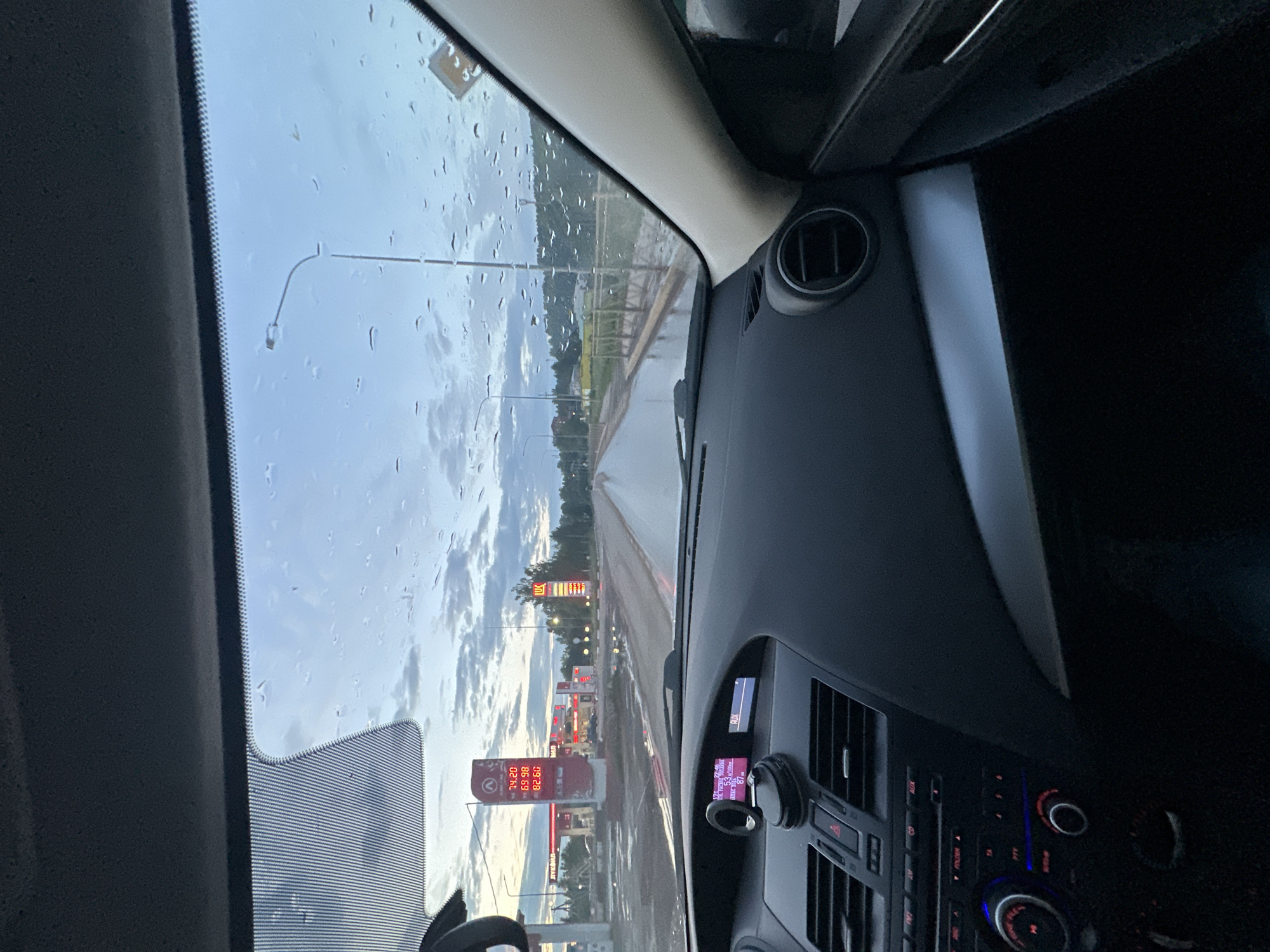 A car's interior view of a road in Ilyinsky, Russia, on a rainy day. The dashboard is visible in the foreground, with a navigation screen displaying "7:57," "17°C," and "53.5 L/100 km," and "AUX." Outside, raindrops are visible on the windshield. A gas station with prominent red signage for "LUX" is to the left, displaying fuel prices of 74.20, 69.98, and 62.60. Streetlights line the wet road, which leads through a semi-urban landscape with trees and buildings in the background. The sky is overcast with clouds.