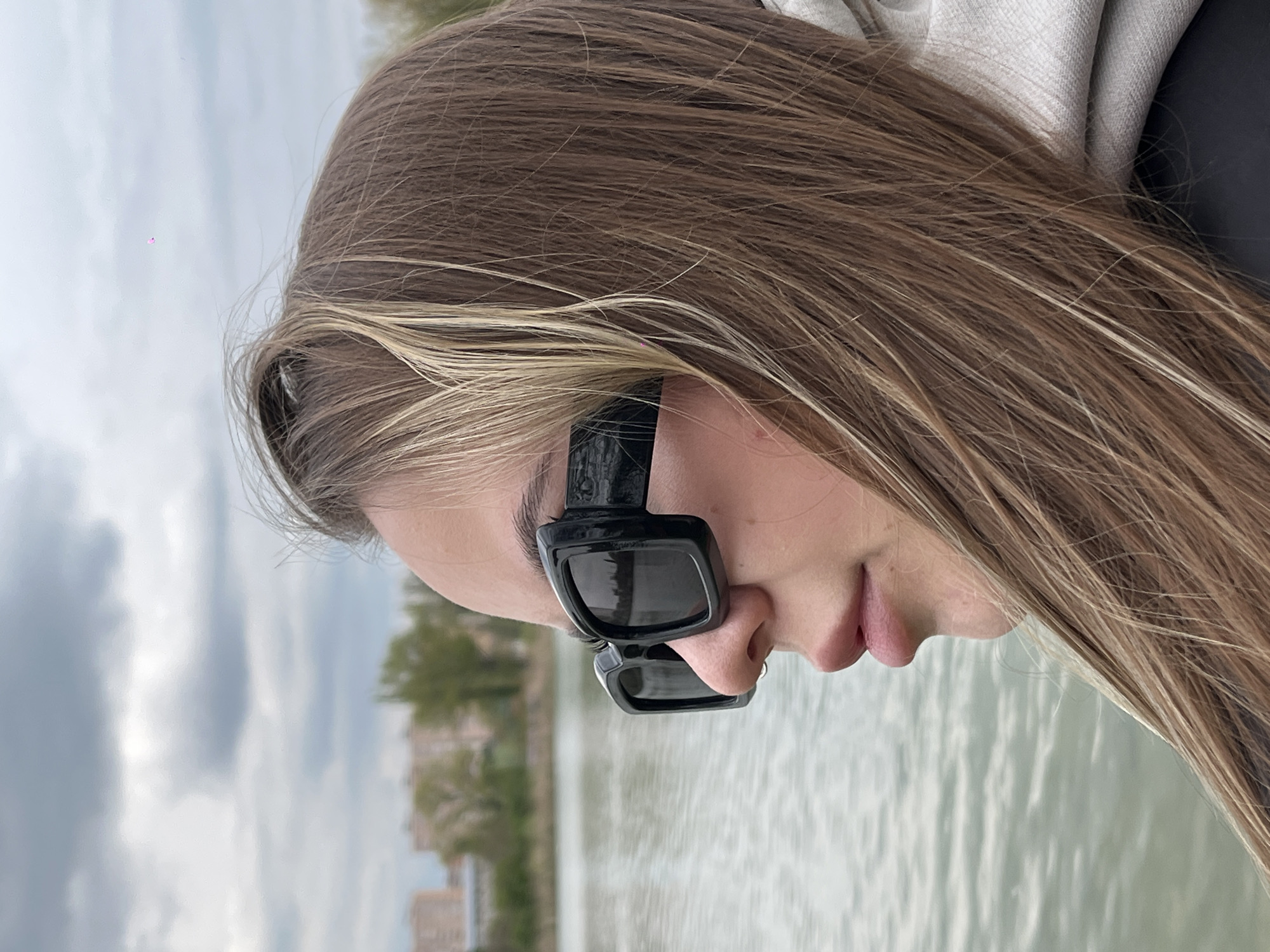 A woman with long, brown hair with blonde highlights wears black rectangular sunglasses and looks down. Behind her is a body of water, likely a river or lake, with trees and buildings visible in the distance. The sky is overcast. The buildings and trees suggest the location is outdoors near a body of water, possibly in a park or urban area. The city of Krasnodar, Russia is not identifiable from this image alone.